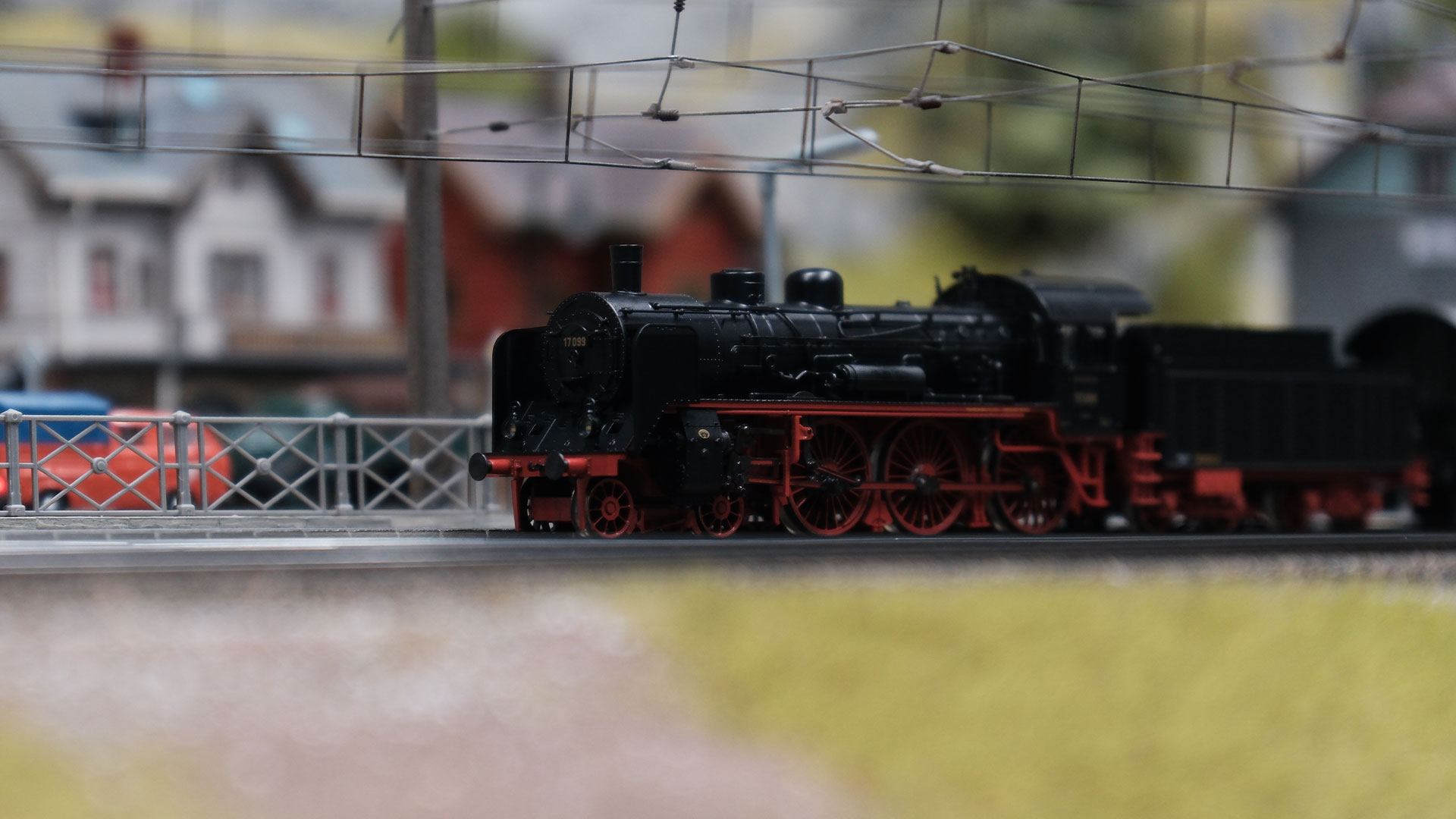 Eine detaillierte Modelldampflokomotive mit roten Rädern steht auf Gleisen in einer Miniaturstadtlandschaft, mit Modellhäusern und einem Zaun im unscharfen Hintergrund.