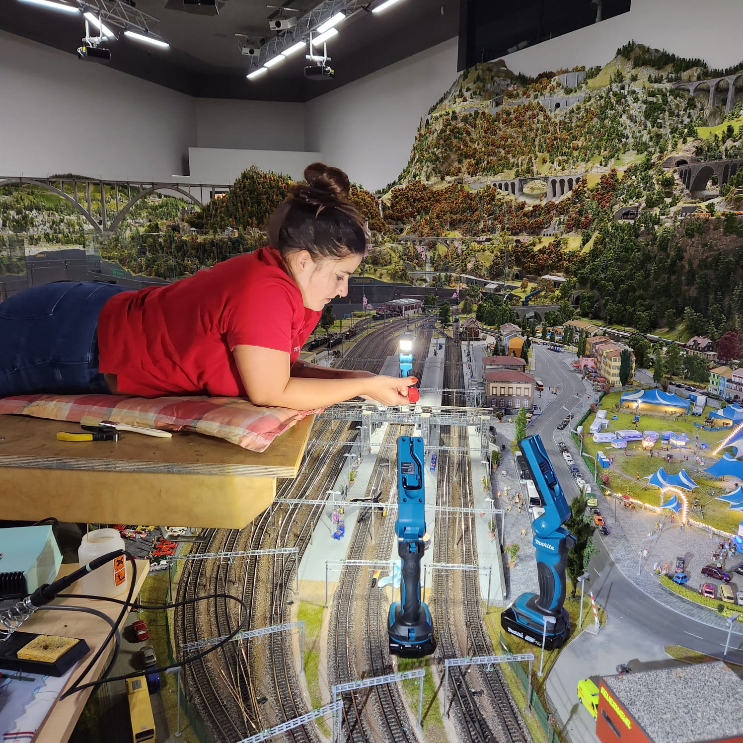Eine Frau in einem roten Hemd liegt auf einem Tisch und arbeitet an einer detailreichen Modelleisenbahn mit Gleisen und Landschaft. Sie benutzt Werkzeuge und betrachtet dabei die Miniaturlandschaft, umgeben von Modelleisenbahnen, Gebäuden und Hügeln.