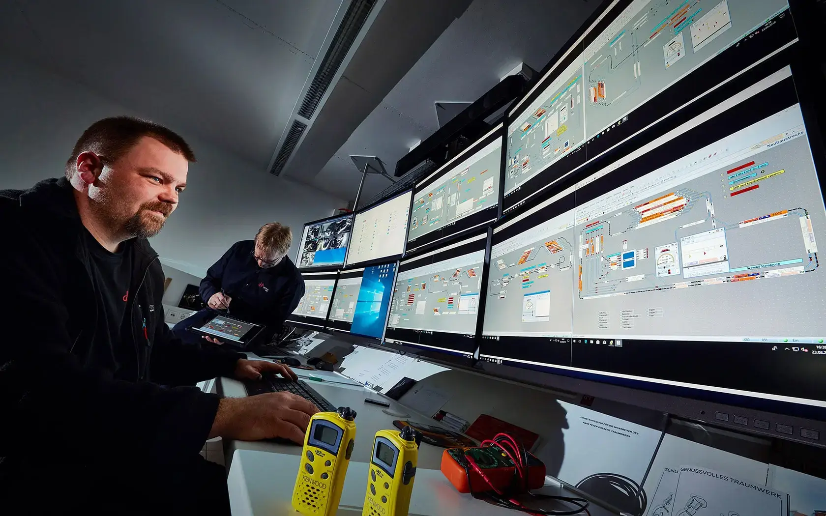 Zwei Männer arbeiten in einem Kontrollraum. Mehrere große Monitore zeigen komplexe Diagramme und Bedienelemente. Ein Mann bedient einen Computer, während der andere Dokumente konsultiert. Im Vordergrund stehen zwei gelbe Funkgeräte auf dem Schreibtisch.