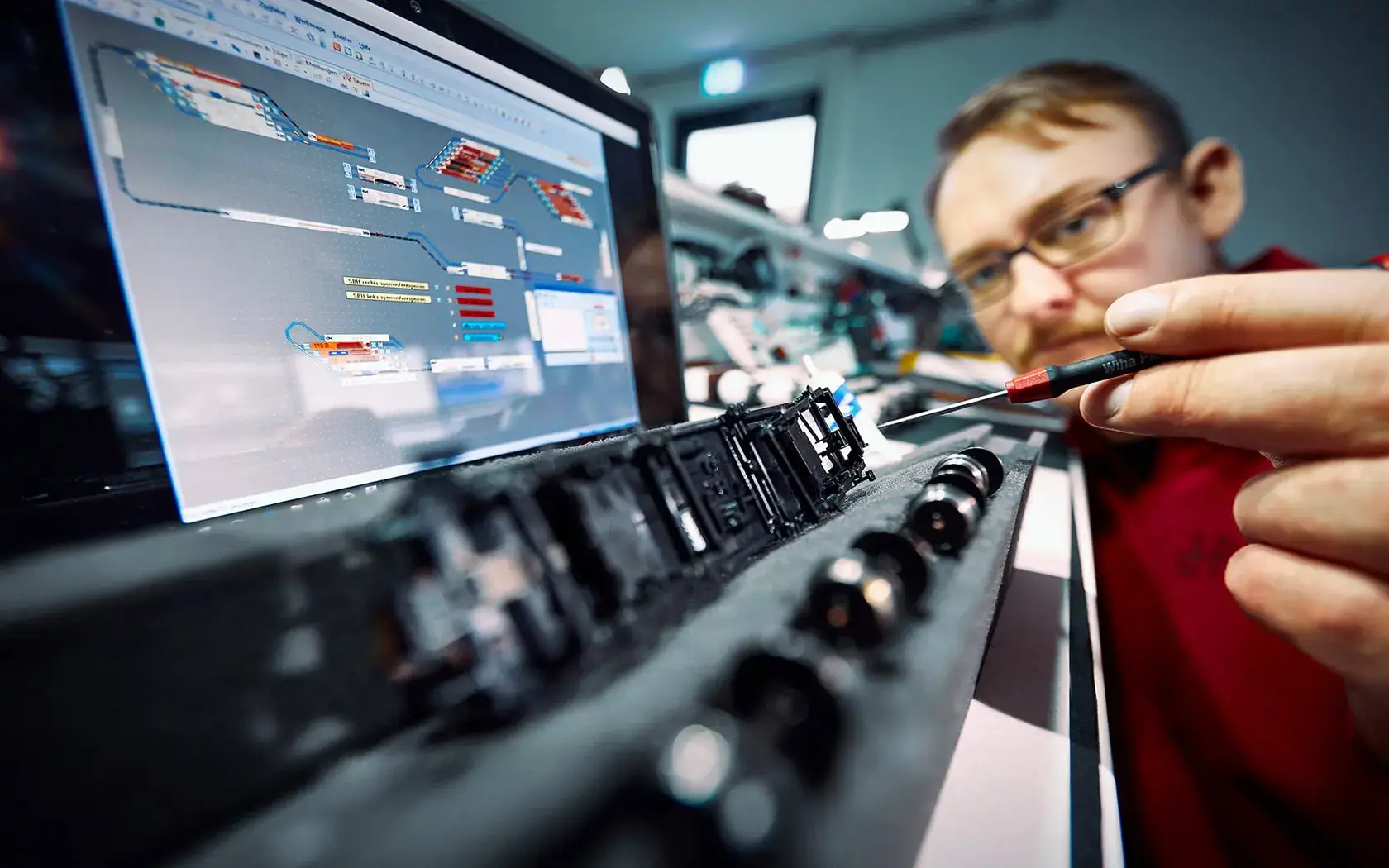 Eine Person bearbeitet mit einem Schraubenzieher eine elektronische Platine. Im Hintergrund zeigt ein Computer ein komplexes Diagramm. Der Fokus liegt auf den Schaltungskomponenten, die Person ist leicht unscharf.