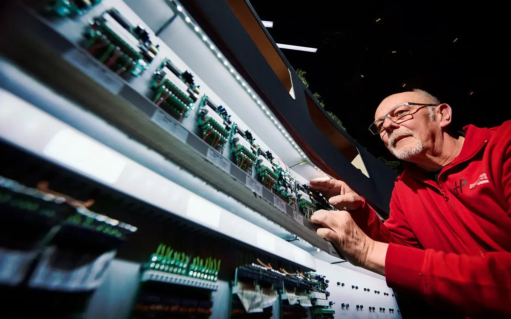 Ein Mann in einem roten Kapuzenpullover justiert oder überprüft Reihen elektronischer Leiterplatten, die auf beleuchteten Regalen in einem Labor oder einer Werkstatt ausgestellt sind.