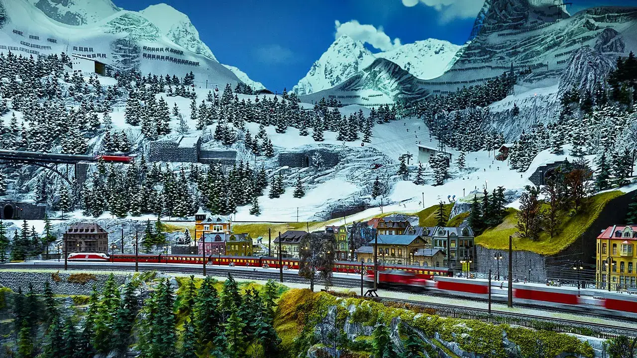Eine detaillierte Modelleisenbahn zeigt rote Züge, die durch ein verschneites Bergdorf mit Kiefern, bunten Gebäuden und hohen, schneebedeckten Gipfeln im Hintergrund fahren.