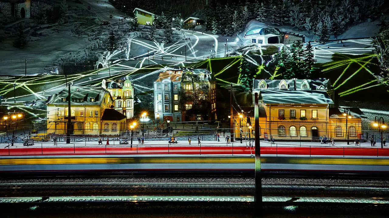 Eine verschneite Dorfszene bei Nacht mit beleuchteten Gebäuden und bunten Lichtspuren, wahrscheinlich von fahrenden Fahrzeugen oder Zügen, mit leuchtenden Linien, die im Hintergrund eine Bergskipiste kreuzen.