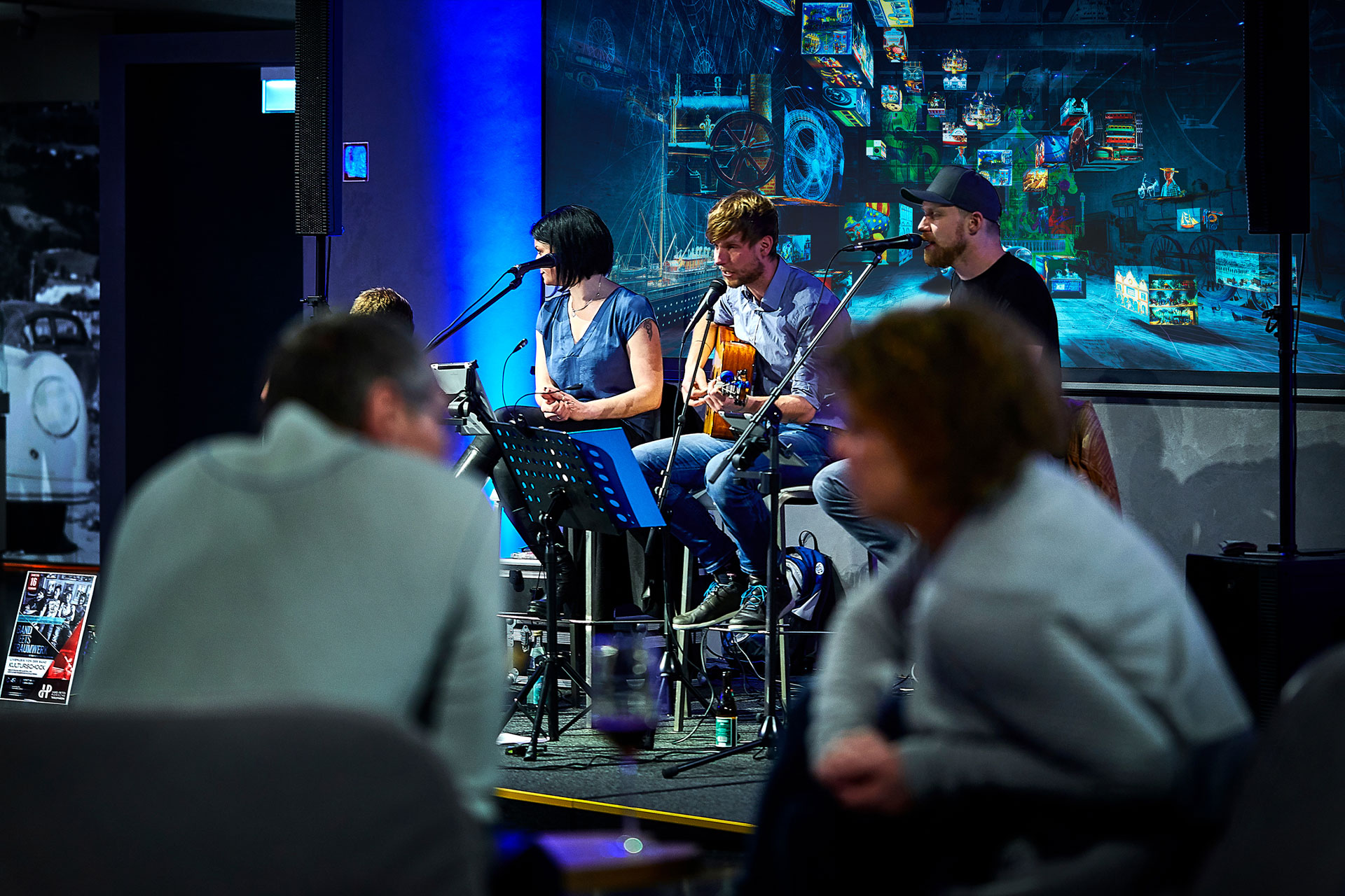 Eine kleine Band spielt live auf der Bühne in einem schwach beleuchteten Raum. Drei Musiker sitzen mit Mikrofonen und Gitarren. Unscharfe Zuschauer sitzen im Vordergrund und hören zu. Hinter der Band befindet sich ein bunter Digitalbildschirm.