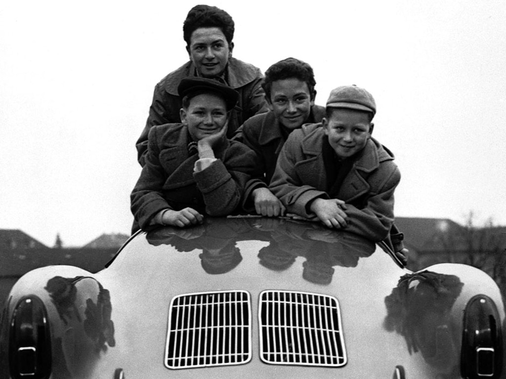 Die vier Söhne von Ferdinand Porsche lehnen sich lächelnd über die Rückbank eines glänzenden Oldtimers. Ihre Spiegelbilder sind auf der Karosserie sichtbar, und im Hintergrund erscheinen die Gebäude verschwommen.