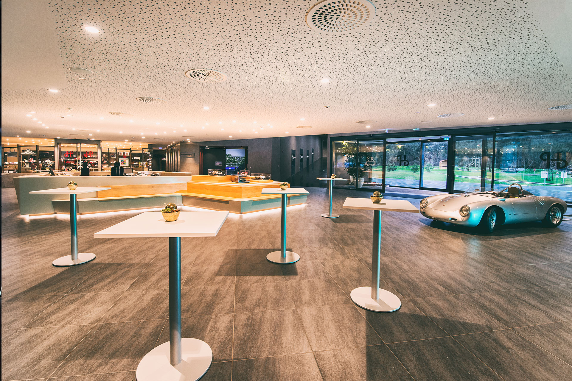 Eine moderne, geräumige Lobby mit glatten Böden, mehreren hohen weißen Tischen, einem hölzernen Empfangstresen und einem klassischen silbernen Cabrio, das neben großen Glasfenstern mit Blick ins Grüne steht.