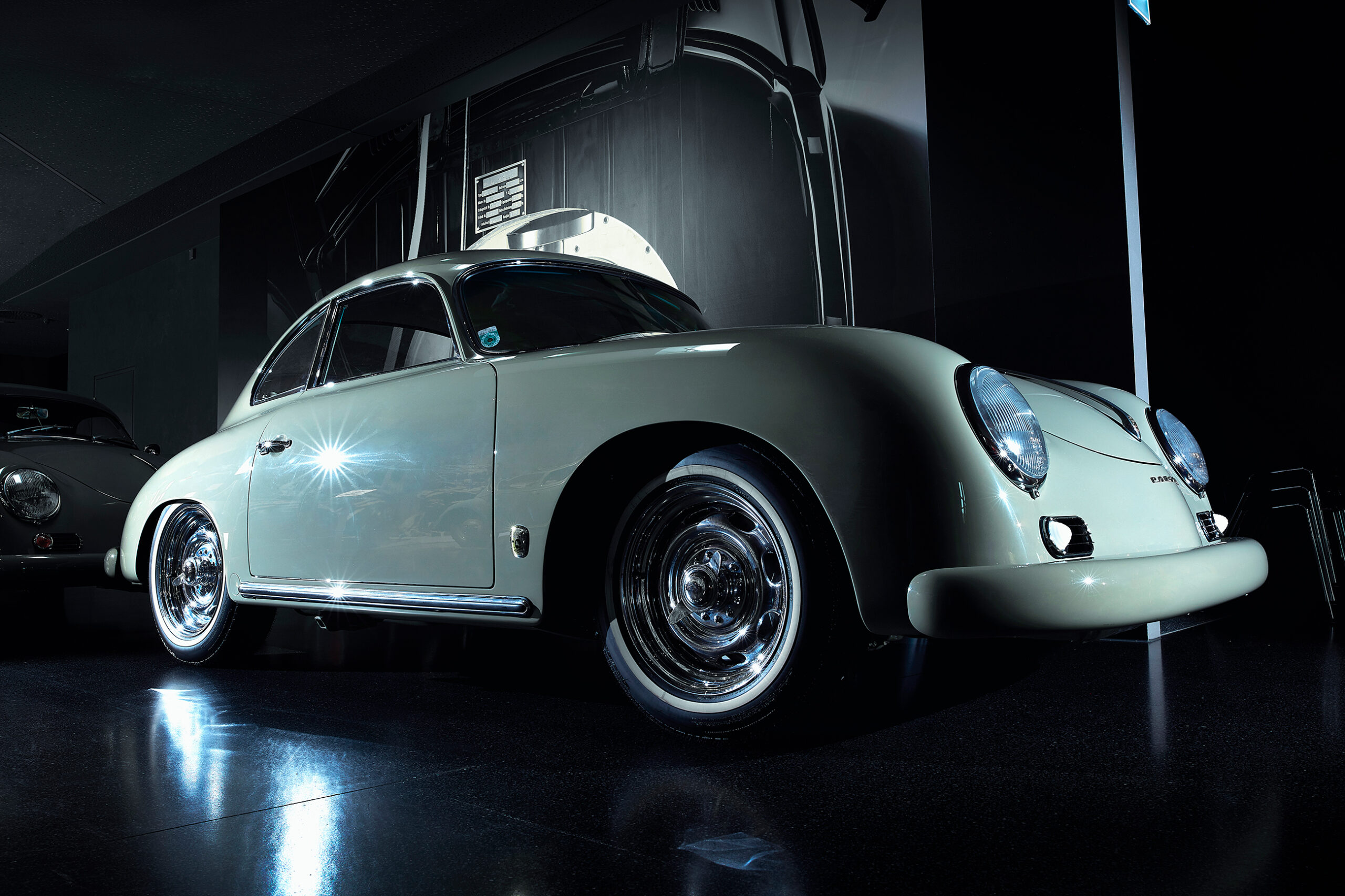 Ein klassisches, glänzend weißes Porsche 356 Coupé wird unter dramatischer Beleuchtung ausgestellt, die seine weichen Kurven und Chromdetails hervorhebt, mit einem dunklen, reflektierenden Boden und einem Oldtimerfoto im Hintergrund.