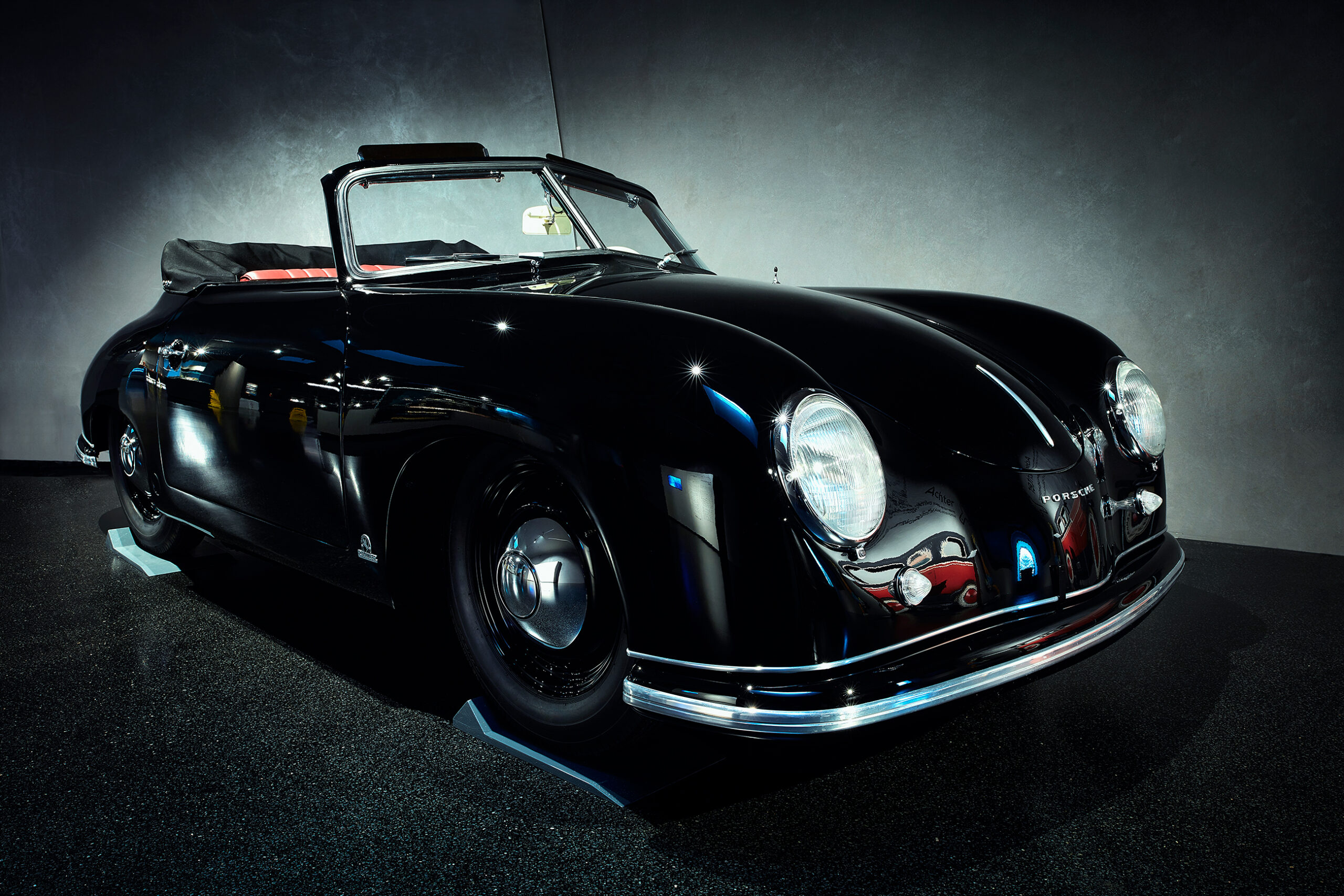 Ein klassisches schwarzes Porsche-Cabriolet mit Stoffverdeck wird im Innenbereich unter dramatischer Beleuchtung ausgestellt, die seine eleganten Kurven und sein Vintage-Design widerspiegelt.
