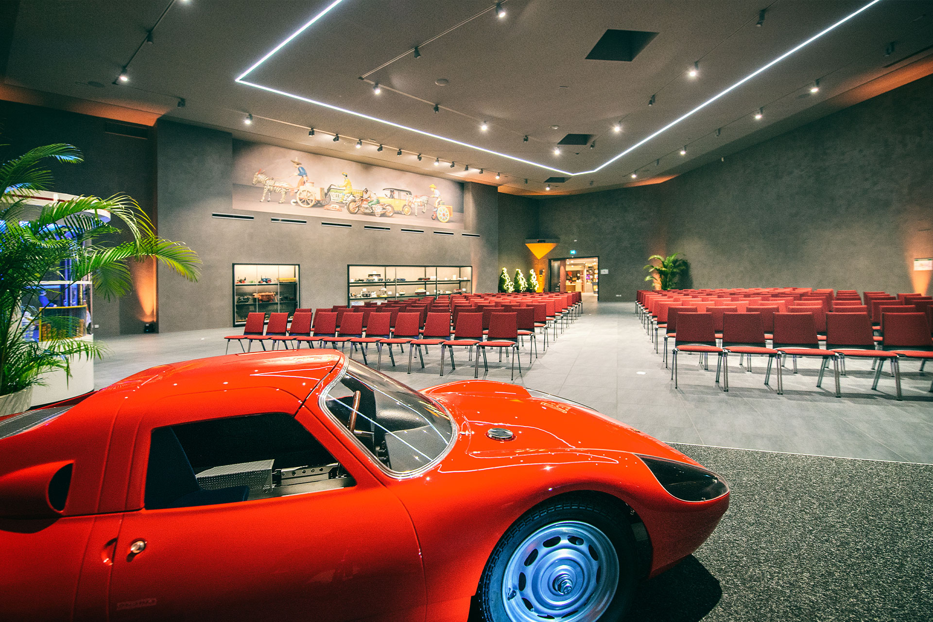 Ein eleganter roter Sportwagen steht im Vordergrund eines modernen, geräumigen Raumes mit Reihen roter Stühle und Autodekor an den Wänden. Der Raum hat graue Wände, helle Beleuchtung und Pflanzen in den Ecken.