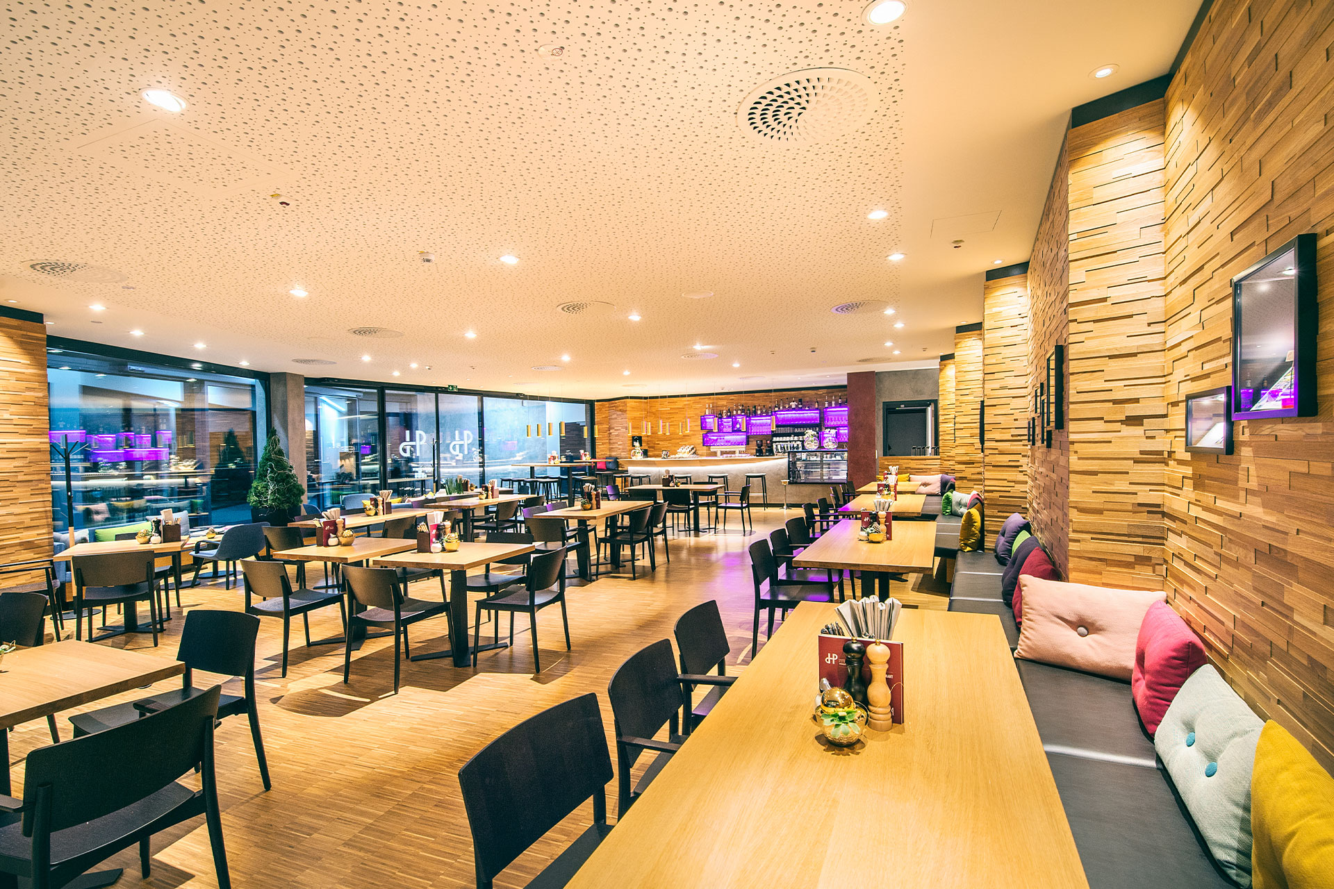 Modernes Restaurant-Interieur mit Wänden und Böden aus Holz, bunten Kissen auf den Sitzbänken, ordentlich angeordneten Tischen und Stühlen und einem gut beleuchteten Barbereich im Hintergrund mit violetter Akzentbeleuchtung.