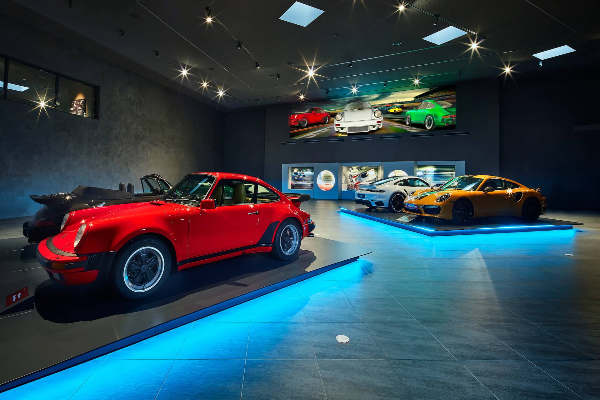 Ein moderner Autosalon mit vier Sportwagen auf beleuchteten Plattformen, darunter ein roter Oldtimer und ein gelbes Auto, mit Autoplakaten an den dunklen Wänden und Scheinwerfern an der Decke.