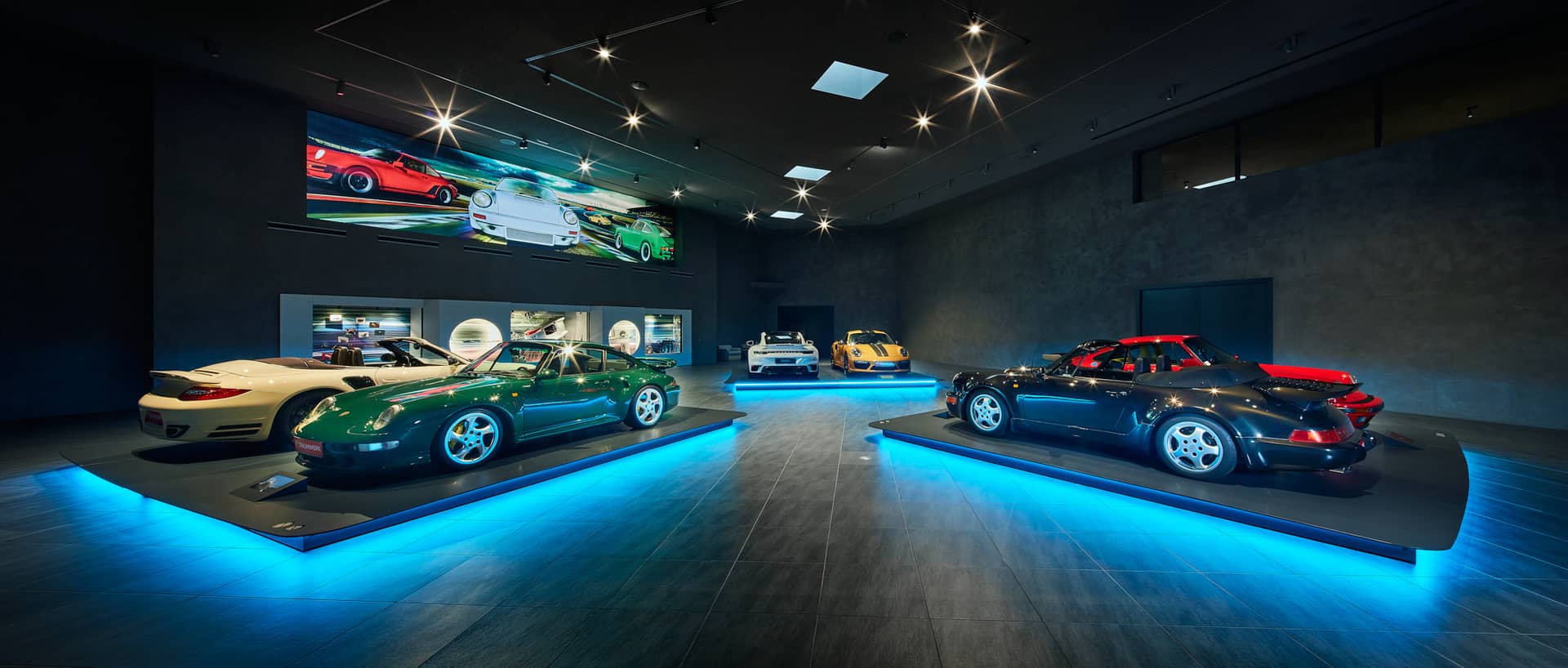 In einem modernen Ausstellungsraum werden mehrere Sportwagen ausgestellt, jeder auf einer blau beleuchteten Plattform. Der dunkle Raum hat glatte, graue Wände, an deren Rückwand ein großes Wandgemälde mit Automobilmotiven hängt.