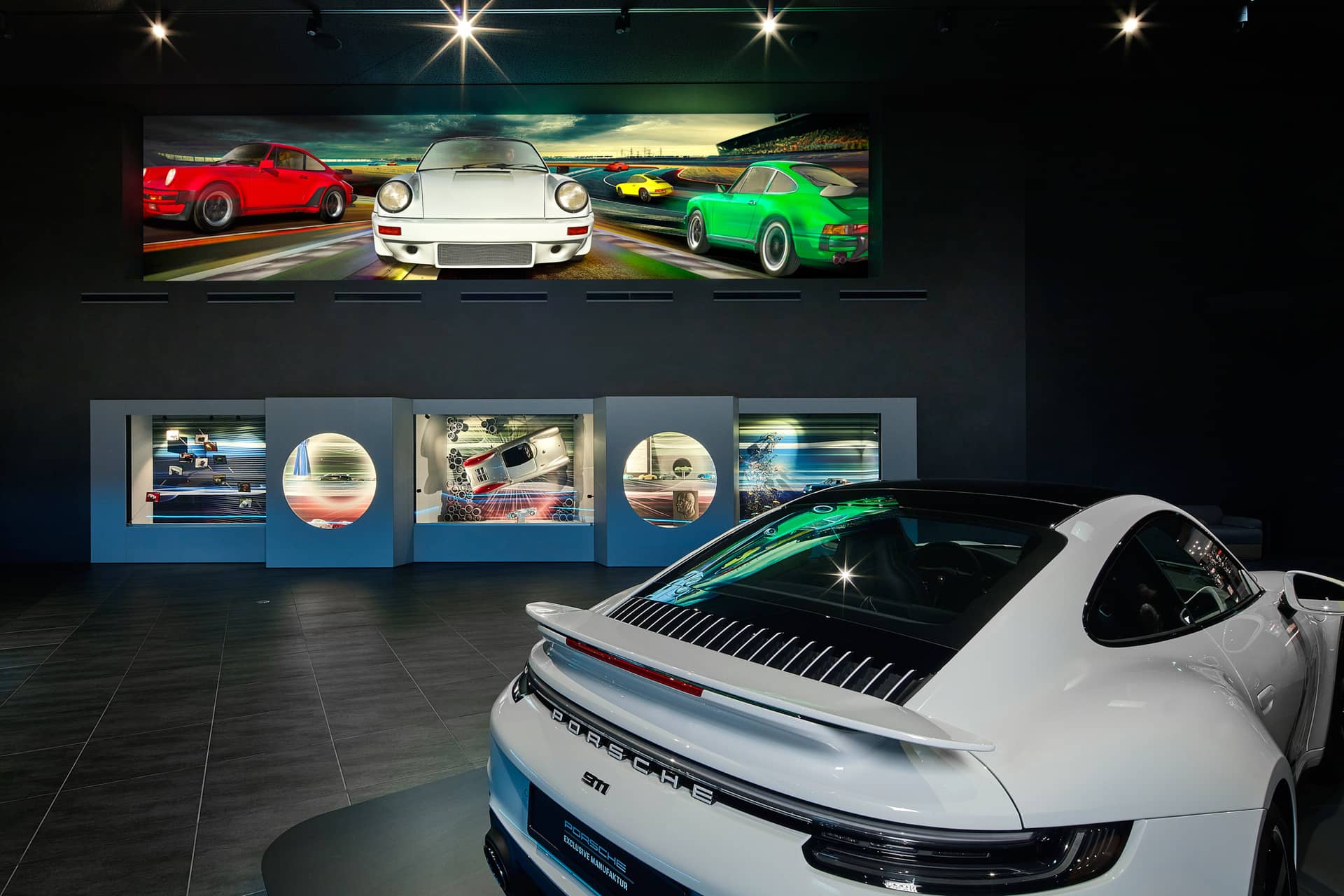Ein weißer Porsche-Sportwagen wird in einem Ausstellungsraum ausgestellt. Darüber ist ein großes Wandbild mit Renn-Porsche-Modellen zu sehen, und in die dunkle Wand dahinter sind mehrere Vitrinen mit Automotiven eingelassen.