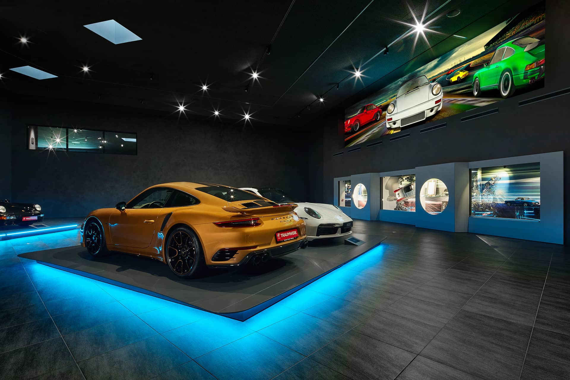 In einem Ausstellungsraum mit moderner Beleuchtung werden auf einer erhöhten Plattform mehrere Luxussportwagen ausgestellt, darunter ein goldenes und ein weißes Auto, mit beleuchteten Wänden und großen Kunstwerken mit Automobilmotiven darüber.