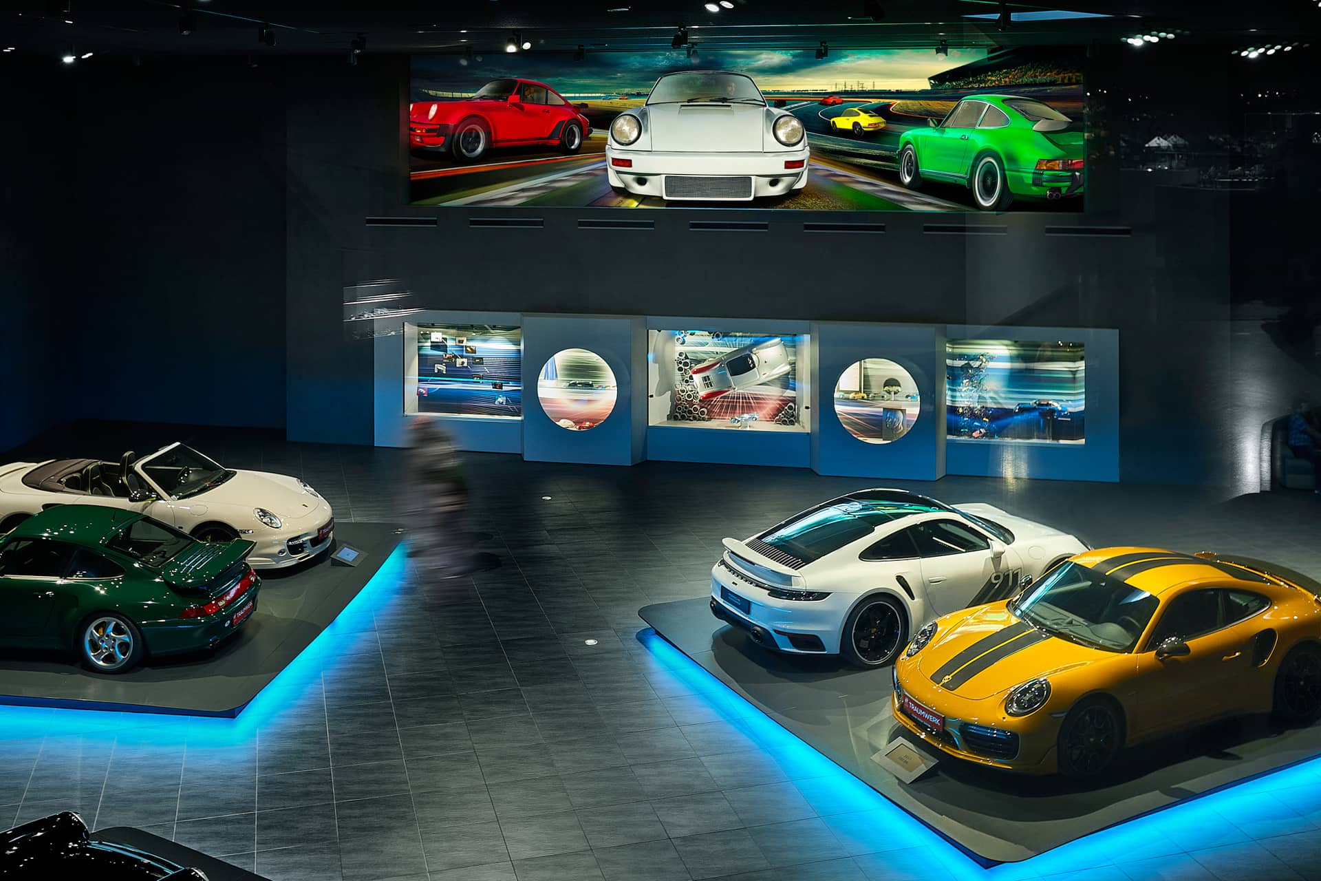 Eine Ausstellung in einem Automuseum mit mehreren Sportwagen, darunter weiße, gelbe und grüne Modelle, mit blauer Bodenbeleuchtung und großen Automobilkunstwerken an der Wand darüber.