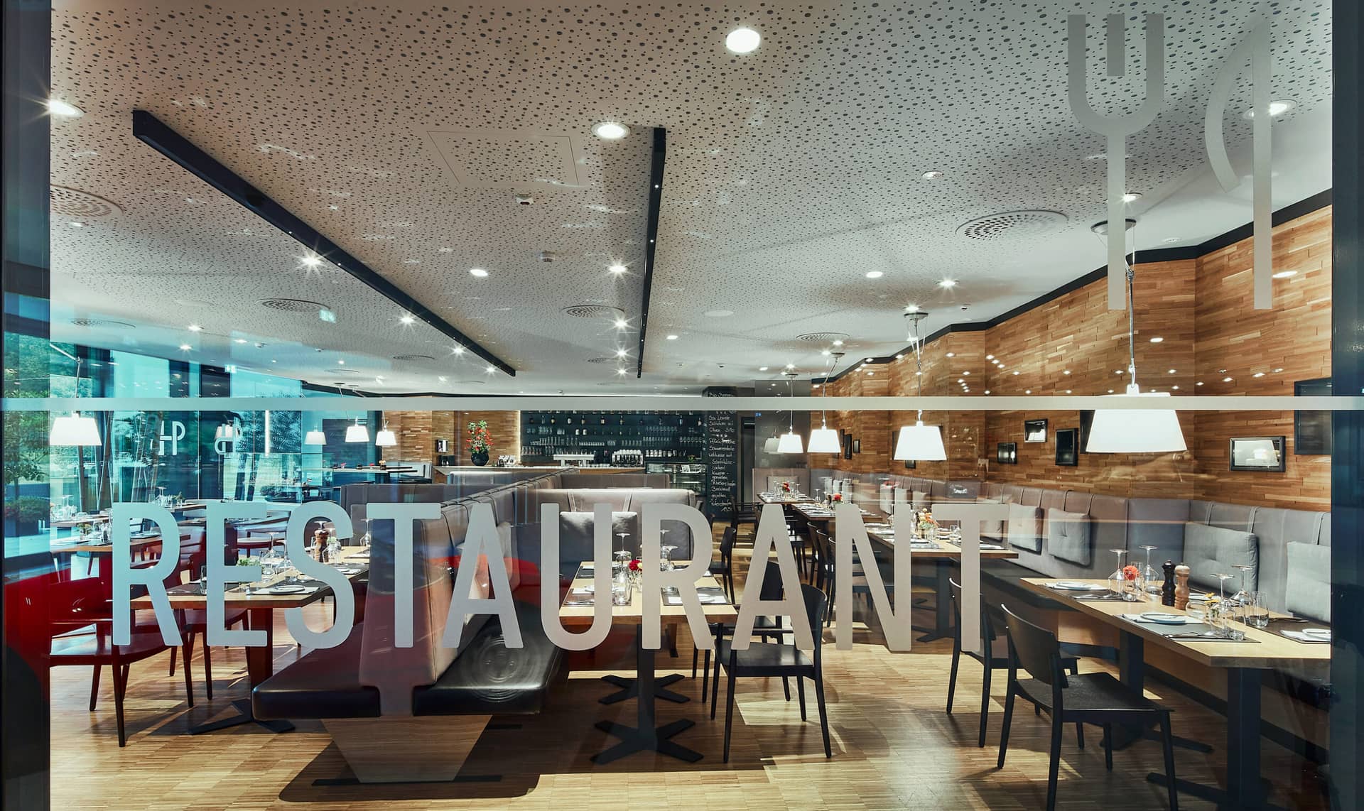 Modernes Restaurant-Interieur mit Holzboden, grauen Sitzecken, gedeckten Tischen und Hängelampen. Das Wort „RESTAURANT“ ist auf die Glasscheibe am Eingang gedruckt. Der Raum wirkt sauber und einladend.