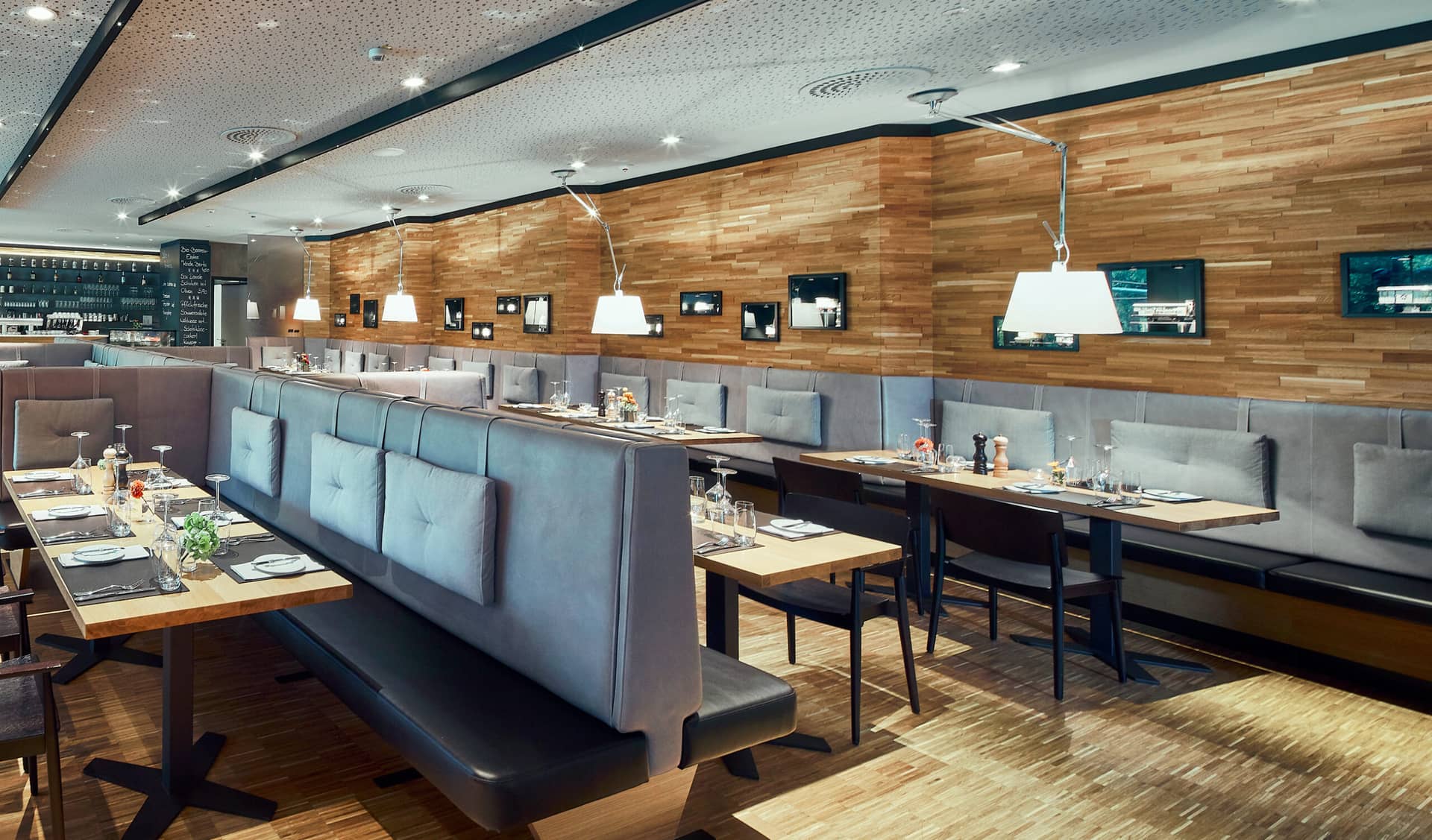 Modernes Restaurantinterieur mit Holzböden und -wänden, grauen gepolsterten Sitznischen, ordentlich eingedeckten Tischen mit Gläsern und Besteck, Hängelampen und gerahmten Fotos an der Wand. Perfekt für eine entspannte Mahlzeit in der Nähe von Traumwerk oder als Ausflugsziel.