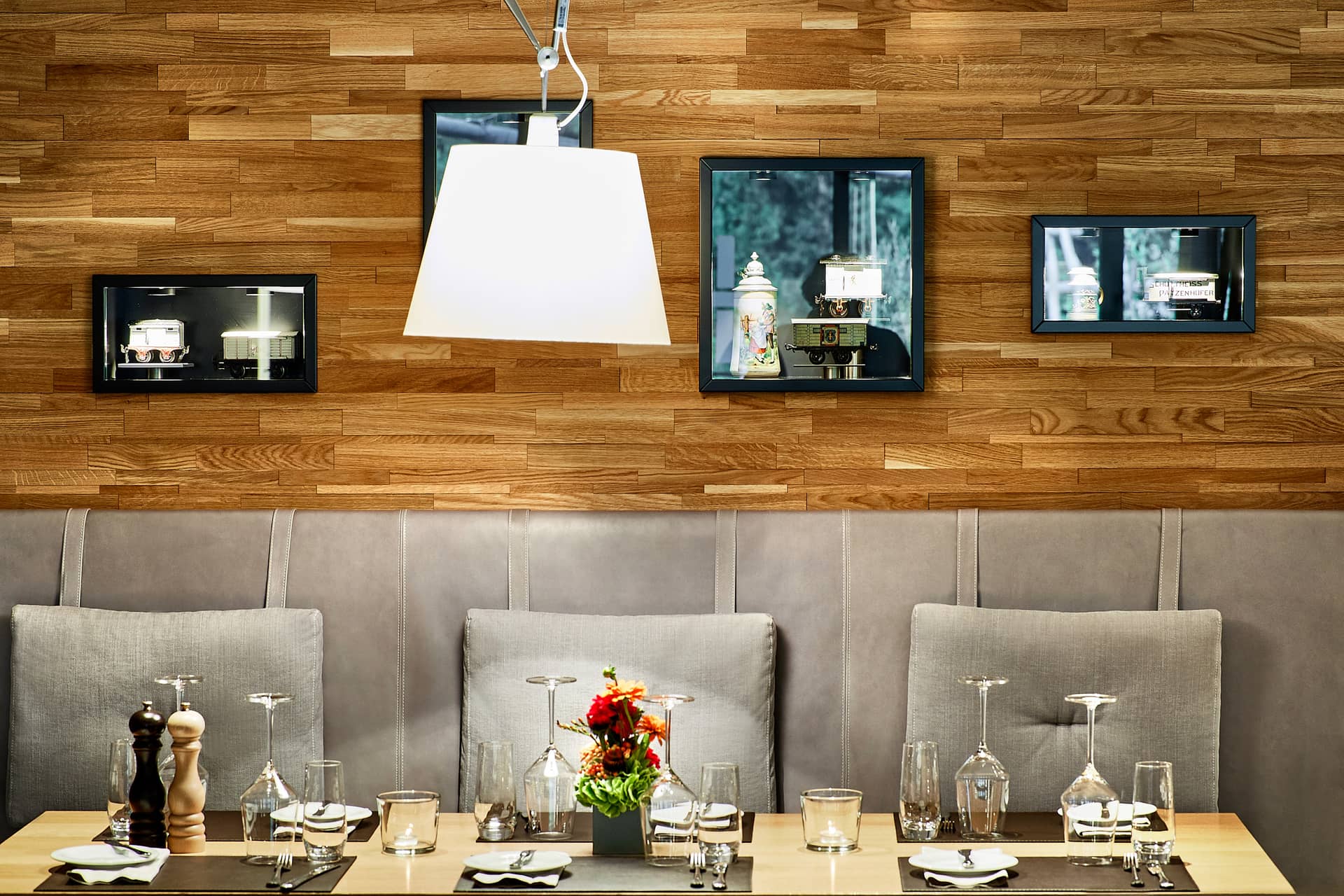 Ein moderner Restauranttisch für vier Personen mit Tellern, Gläsern, Besteck und einem kleinen Blumenarrangement vor einer Holzwand mit grauen Kissen und dekorativen Regalen, auf denen Teekannen und andere Gegenstände ausgestellt sind.