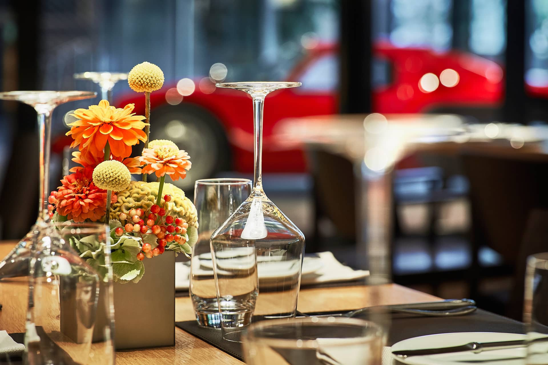 Nahaufnahme eines mit Gläsern, Tellern und orangefarbenen Blumen gedeckten Tisches in einem Restaurant; ein roter Porsche steht unscharf im Hintergrund vor dem Fenster.