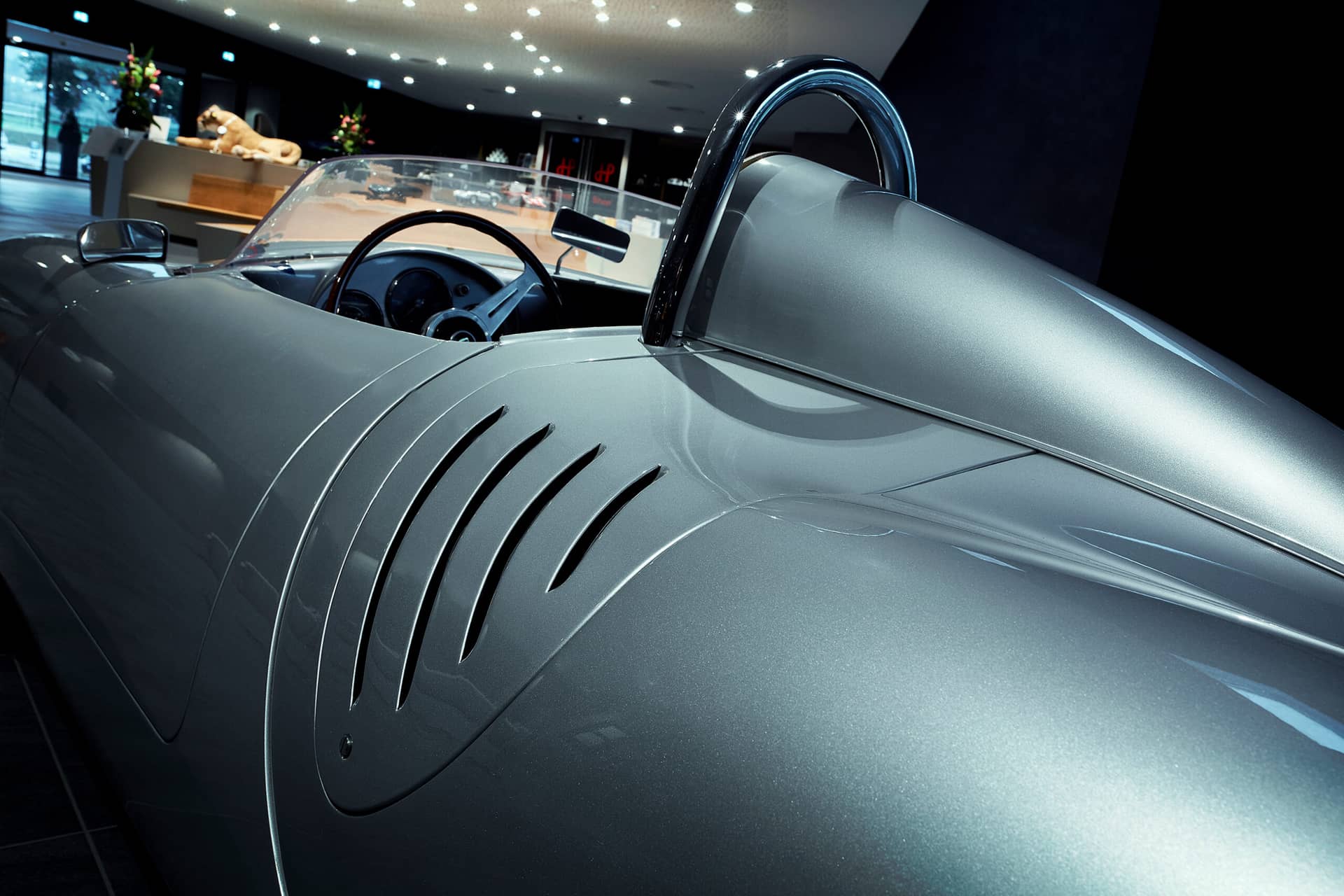 Nahaufnahme eines klassischen silbernen Sportwagens mit Fokus auf die geschwungene Karosserie und das Cockpit mit sichtbarem Lenkrad im Inneren. Das Auto wird im Innenbereich ausgestellt, im Hintergrund sind weitere Fahrzeuge und Dekorationen zu sehen.