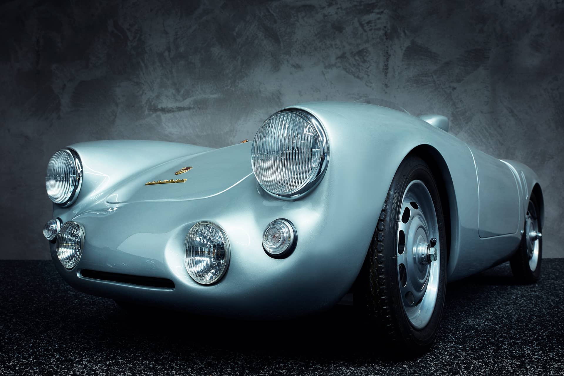 Eine Untersicht eines klassischen silbernen Porsche-Sportwagens mit runden Scheinwerfern und einem eleganten Vintage-Design, der in einem Innenbereich vor einer strukturierten grauen Wand geparkt ist.