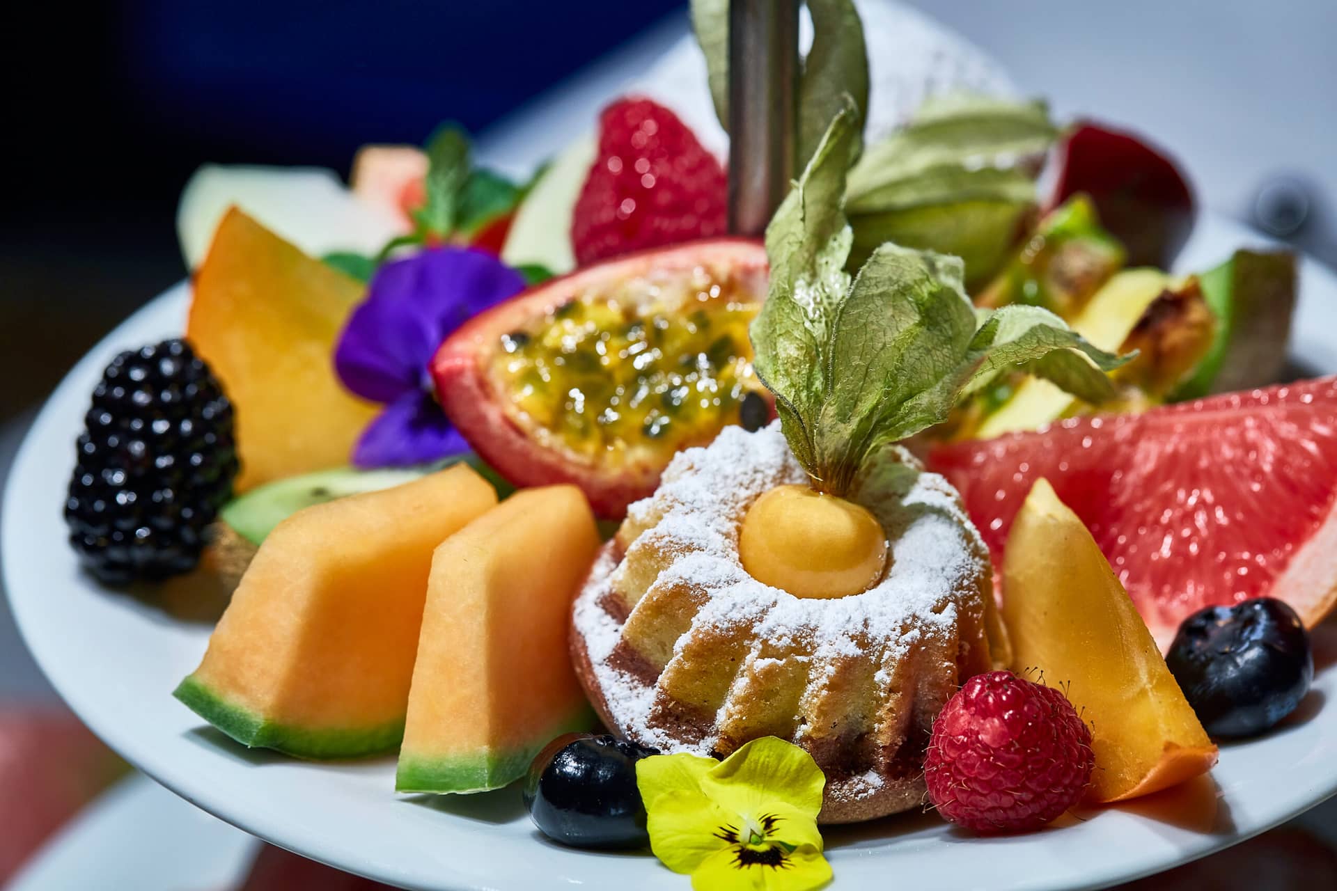 Ein Teller mit verschiedenen frischen Früchten, essbaren Blumen und einem kleinen, mit Puderzucker bestäubten Kuchen, umgeben von Beeren, Melonenscheiben, Passionsfrüchten und bunten Garnierungen - perfekt, um ihn nach der Erkundung des Traumwerks als Ausflugsziel zu genießen.
