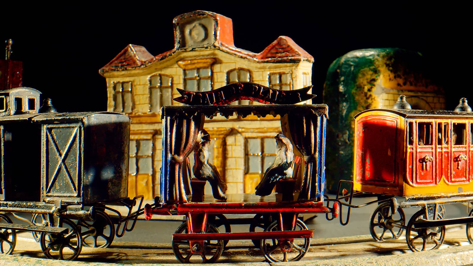 Miniatur-Spielzeugeisenbahnwaggons und eine kleine Bühne mit zwei Figuren stehen vor einem Modellgebäude, das einem Bahnhof ähnelt. Alles ist in leuchtenden Farben bemalt und mit detaillierten Details versehen, unter dramatischer Beleuchtung.