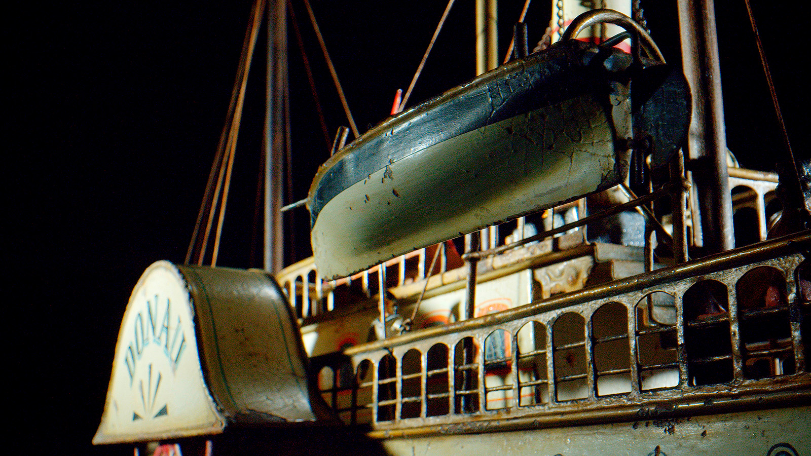 Nahaufnahme eines alten, detaillierten Modelldampfschiffs mit einem an Davits hängenden Rettungsboot, verzierten Fenstern und einem großen Schaufelrad, alles mit einem verwitterten Vintage-Aussehen vor einem dunklen Hintergrund.