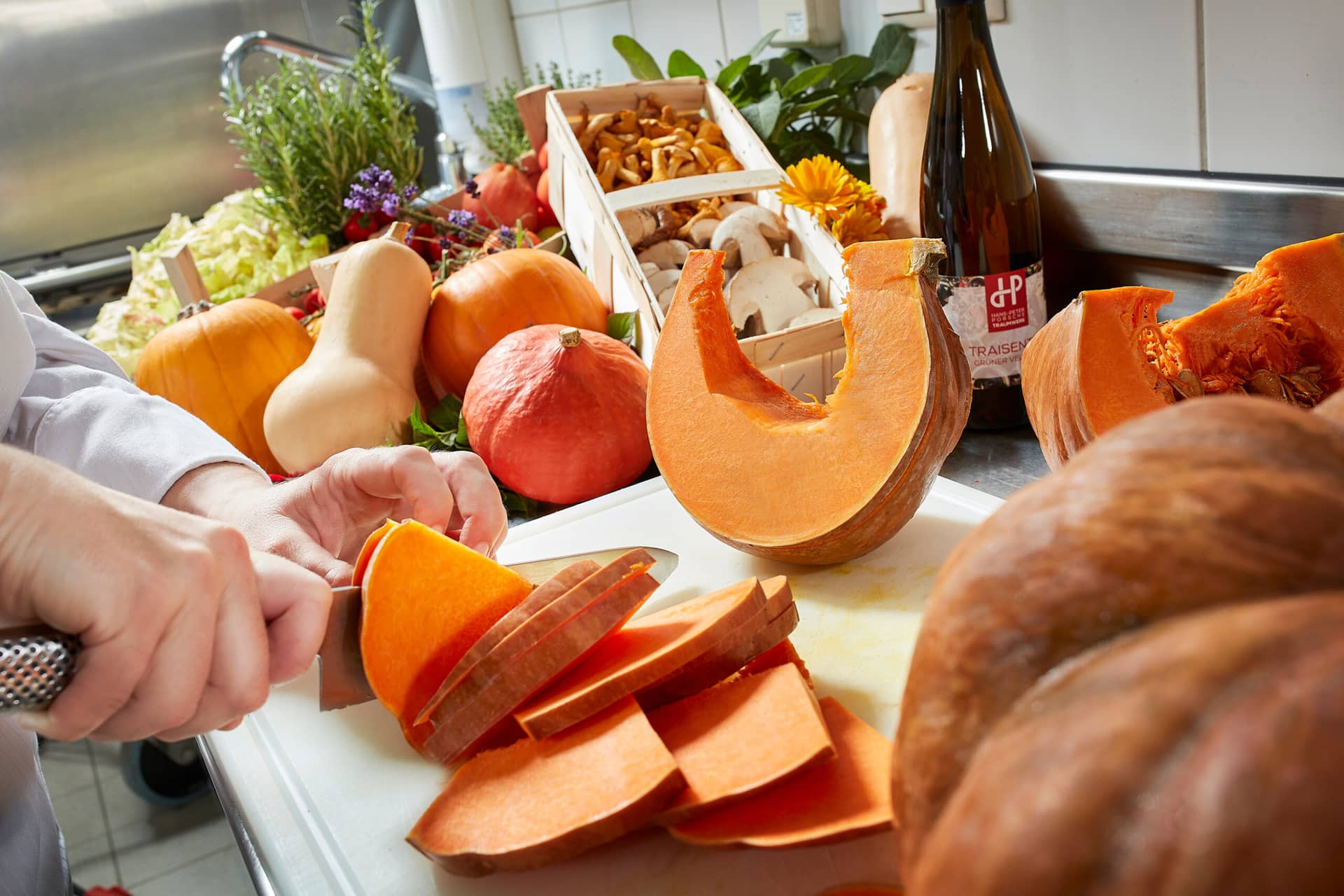 Eine Person schneidet einen Kürbis auf einem Schneidebrett in einer Küche, umgeben von porscheförmigen Kürbissen, Pilzen, frischen Kräutern und einer Flasche Wein. Der Arbeitsbereich ist mit farbenfrohem Herbstgemüse gefüllt und hat eine gemütliche Traumwerk-Atmosphäre.