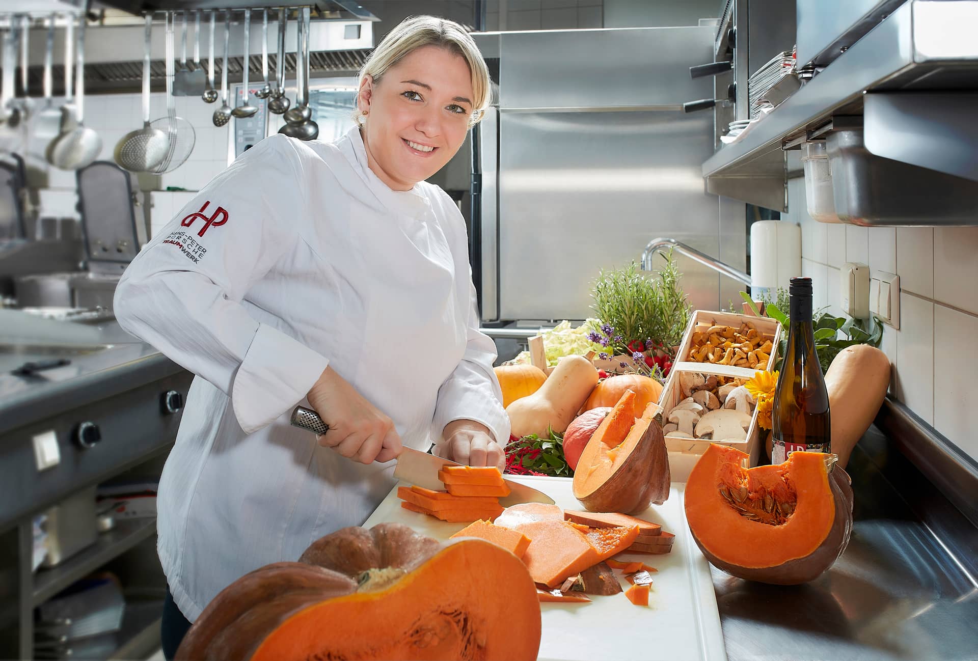 Ein Koch in weißer Uniform lächelt, während er in einer professionellen Edelstahlküche einen großen orangefarbenen Kürbis auf einer Küchentheke schneidet, die mit verschiedenen Gemüsesorten, Pilzen und einer Flasche gefüllt ist.