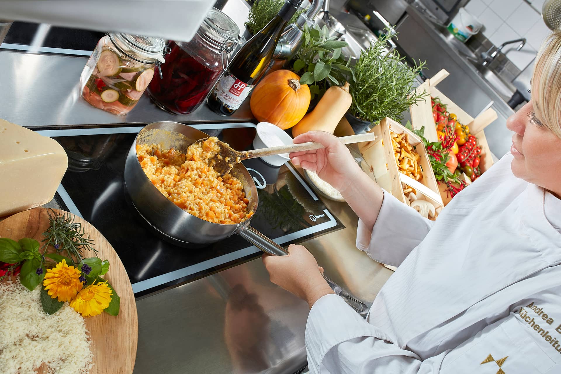 Ein Koch in weißer Uniform rührt in einer Küche des Traumwerks einen Topf mit Risotto, umgeben von frischen Kräutern, Gemüse, Käse, Wein und Gläsern mit eingelegtem Gemüse - ein kulinarisches Erlebnis, das zum idealen Ausflugsziel eines jeden Porsche-Liebhabers passt.
