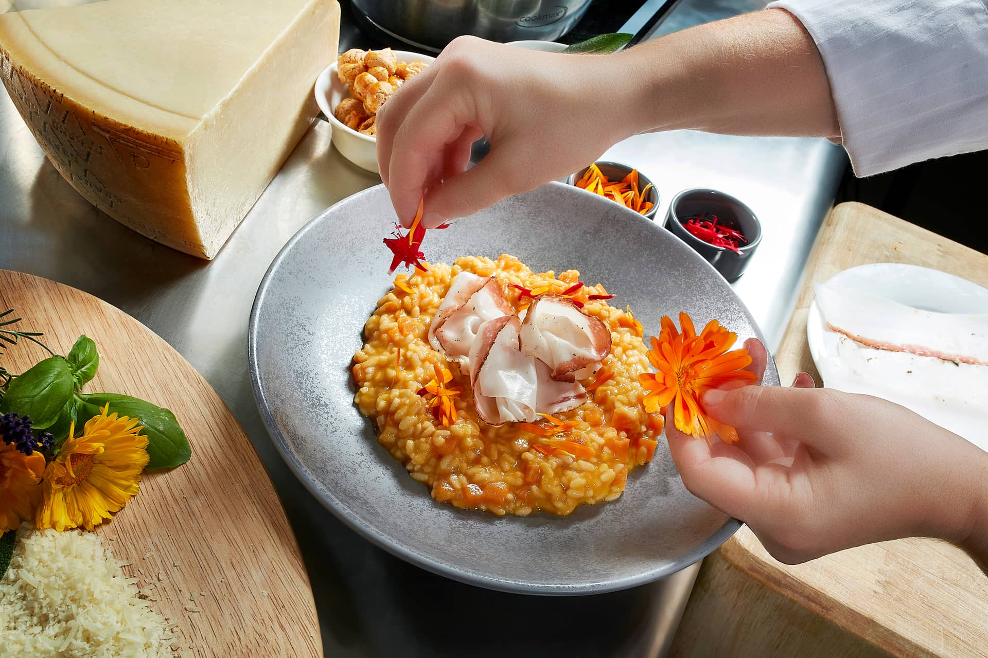 Ein Koch garniert einen Teller mit cremigem Risotto mit essbaren Blüten und dünnen Fleischscheiben, umgeben von Käse, Kräutern, Safran und Küchenutensilien auf einer Arbeitsplatte aus Edelstahl im renommierten Traumwerk - ein perfektes Ausflugsziel für Feinschmecker.