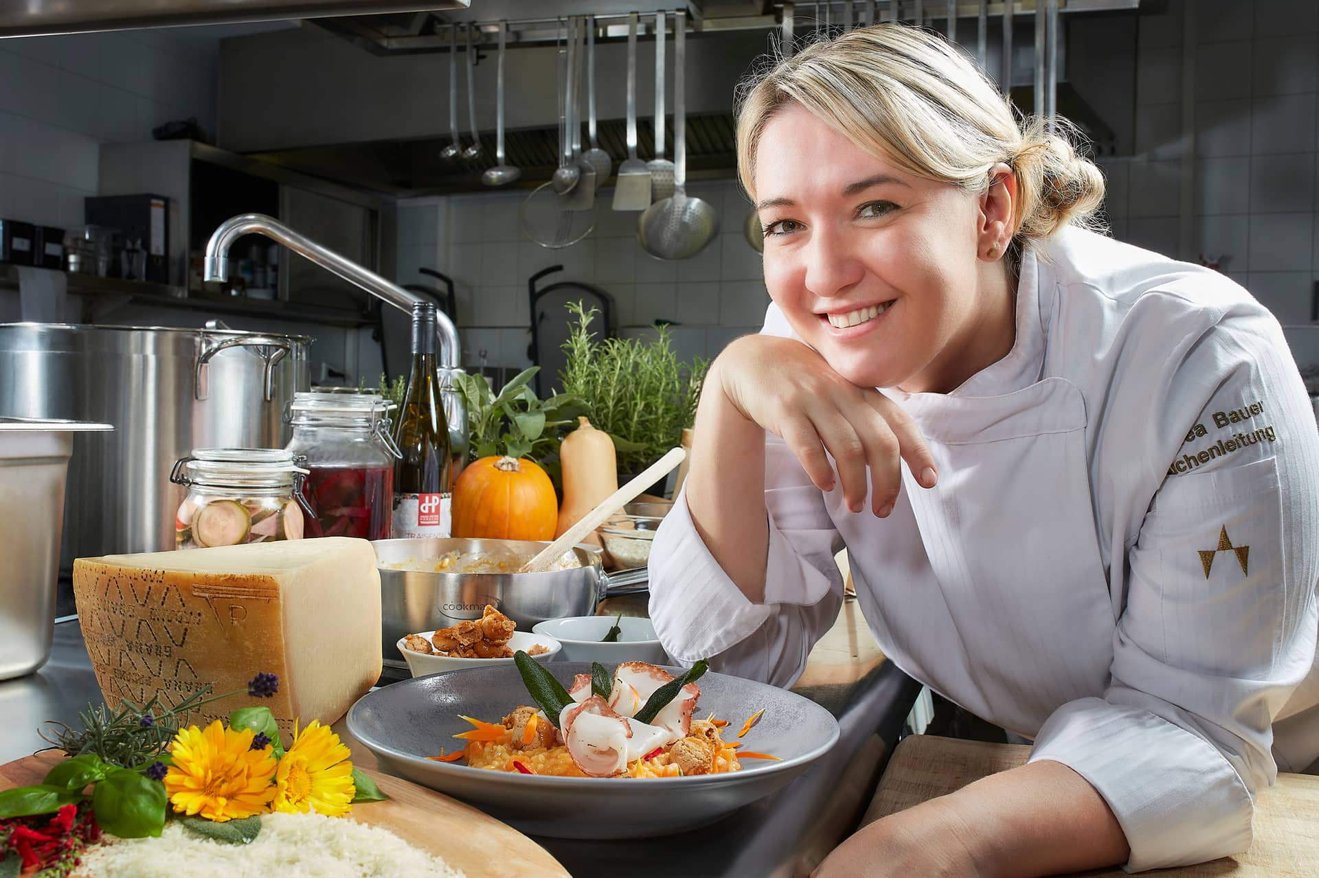 Ein lächelnder Koch in weißer Uniform lehnt an einer Küchentheke im Traumwerk, neben einem aufgetischten Gourmetgericht, Kräutern, Käse und Blumen, mit verschiedenen Küchengeräten und Zutaten im Hintergrund - ein perfektes Ausflugsziel für Feinschmecker.