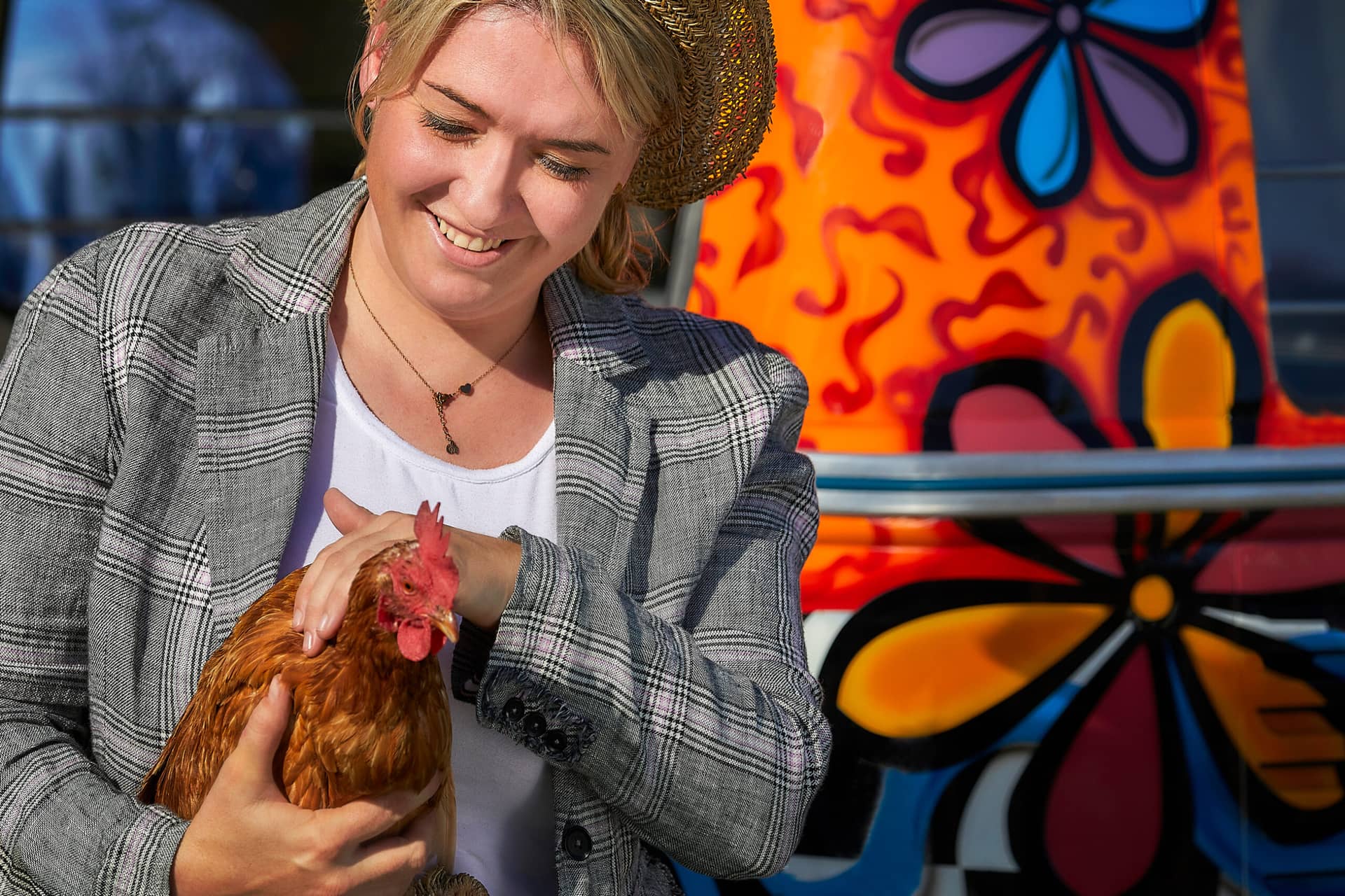 Eine lächelnde Person in einem karierten Blazer und einem Strohhut hält sanft ein braunes Huhn. Dahinter steht ein bunt bemaltes Fahrzeug mit farbenfrohen Blumenmustern in Orange, Rosa, Gelb und Blau - perfekt für ein traumwerk-inspiriertes Ausflugsziel.