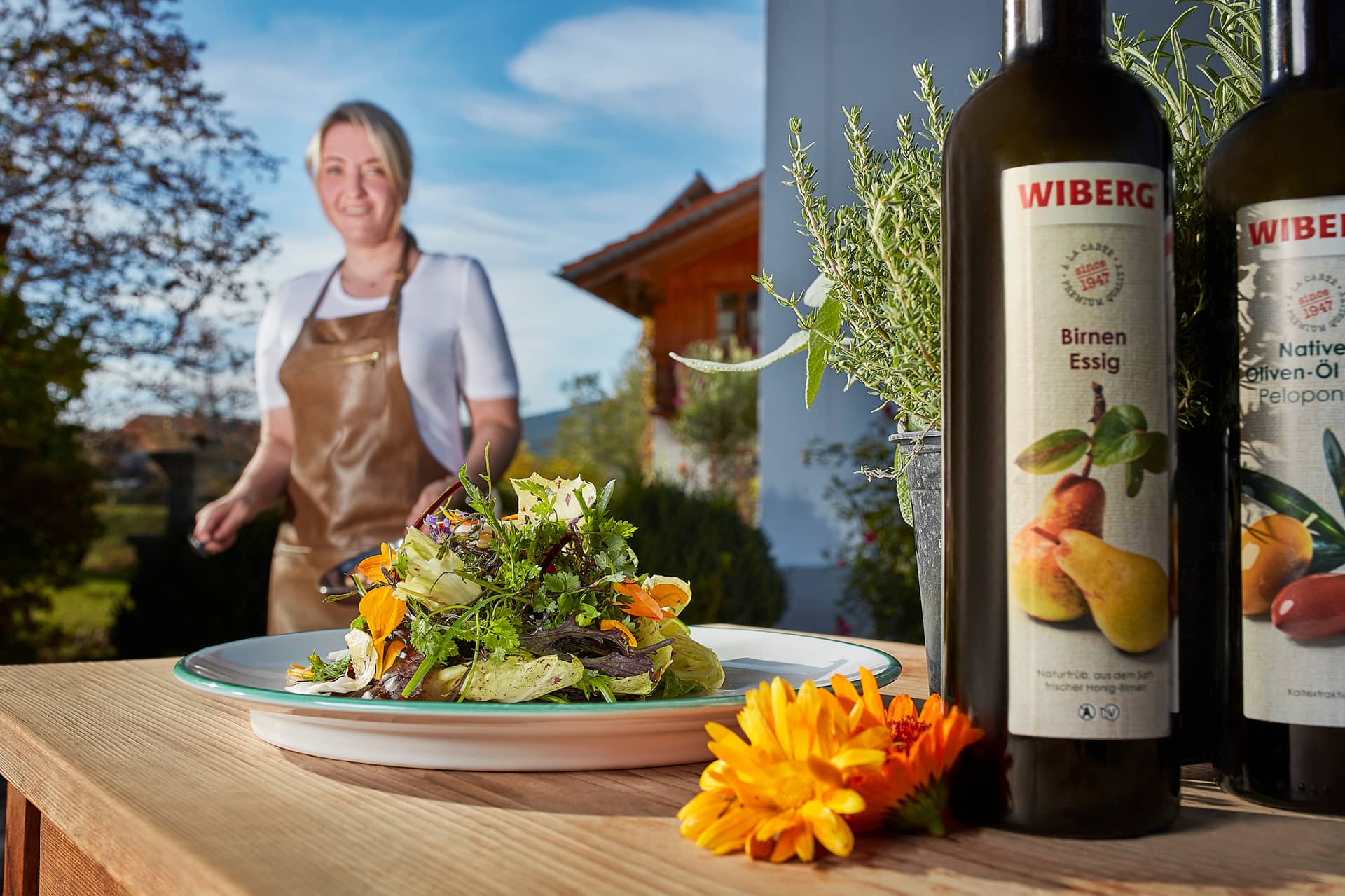 Ein bunter Salat auf einem Teller liegt auf einem Holztisch neben einer Ringelblume und Flaschen mit Wiberg-Birnenessig und Olivenöl. Im Hintergrund lächelt eine Frau in einer Schürze - perfekt für ein ausflugsziel Mittagessen im Freien.