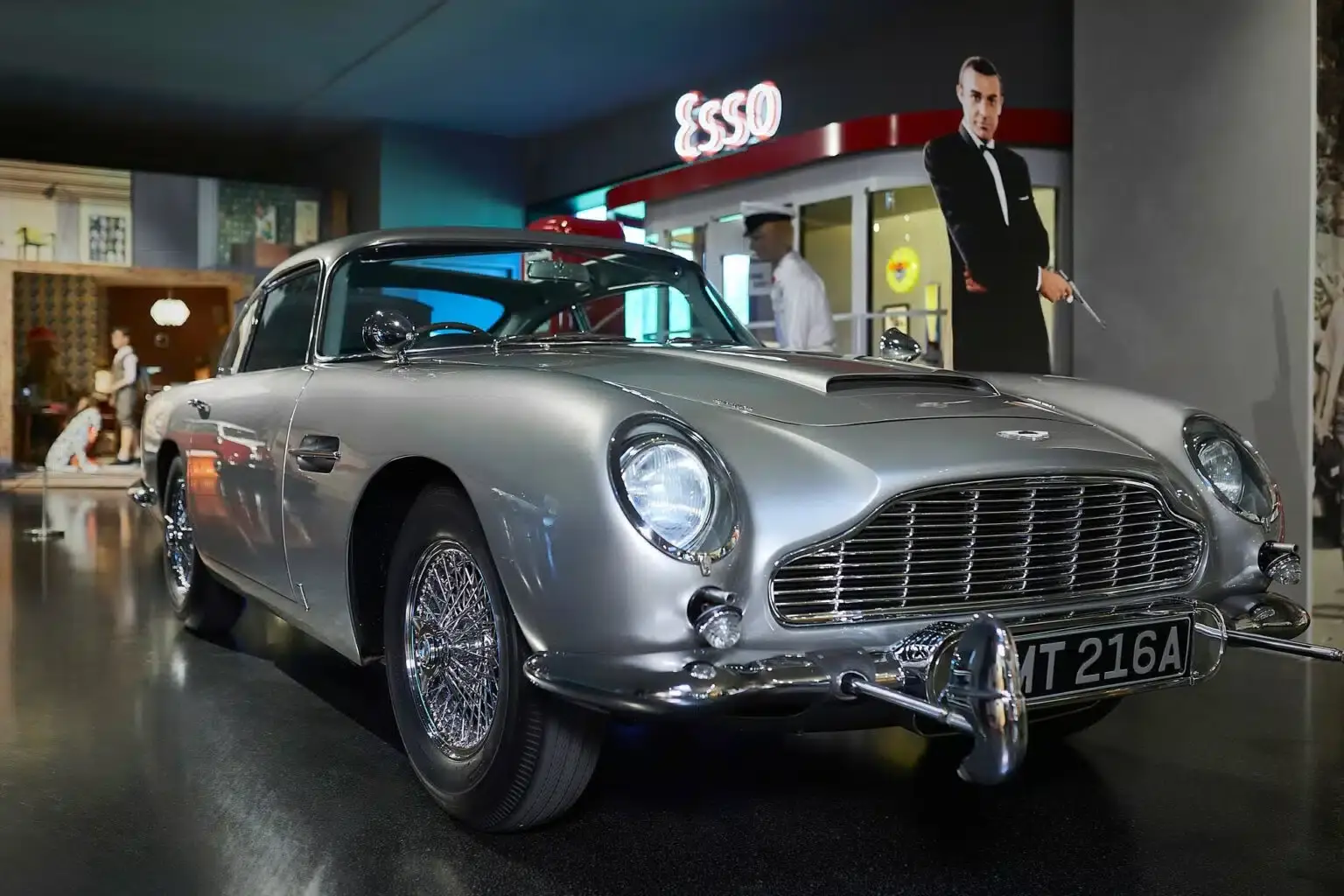 Im Innenbereich steht ein klassischer silberner Aston Martin mit dem Kennzeichen „NT 216A“. Dahinter sind ein Esso-Schild und der lebensgroße Ausschnitt eines Mannes im Smoking zu sehen, der wahrscheinlich auf ein James-Bond-Thema verweist.