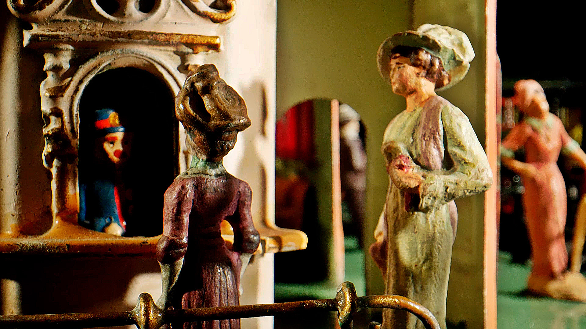 Zwei bemalte Frauenfiguren stehen neben einem Ticketschalter, in dem eine uniformierte Gestalt sitzt. Die Szene wirkt altmodisch oder wie ein Miniaturdiorama mit detailreicher zeitgenössischer Kleidung und architektonischen Elementen.
