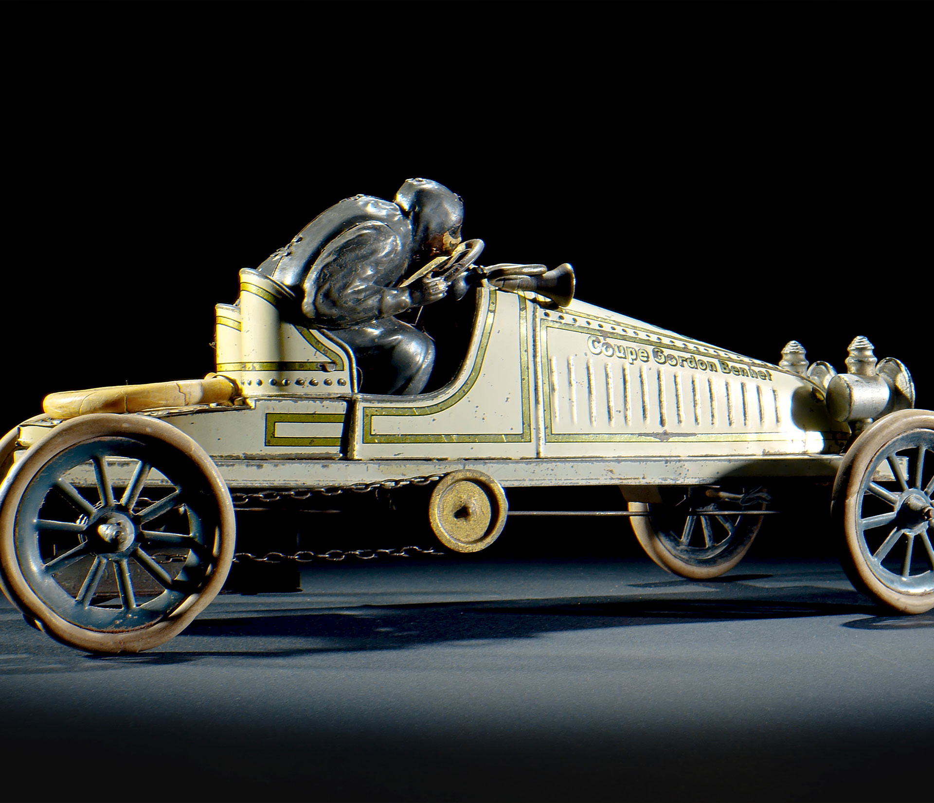 Ein Oldtimer-Spielzeugauto mit sitzendem Fahrer, weiß lackiert mit goldenen Akzenten, mit großen Speichenrädern und detaillierten Gravuren. Das Auto hat die Aufschrift George Gordon Bennett auf der Seite - perfekt für jeden Porsche Traumwerk Sammler.