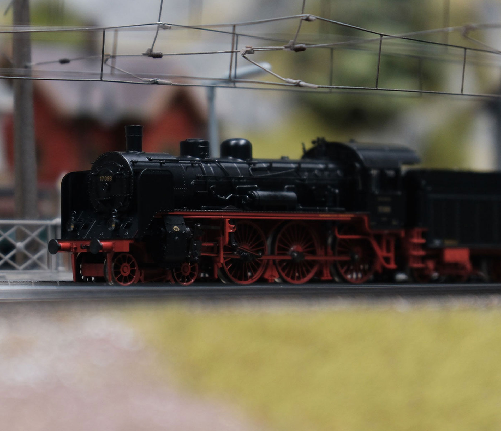 Ein detailliertes Modell einer alten schwarzen Dampflokomotive mit roten Rädern steht auf einem Gleis im Traumwerk, einem idealen Ausflugsziel, mit verschwommener Landschaft und Oberleitungen im Hintergrund.
