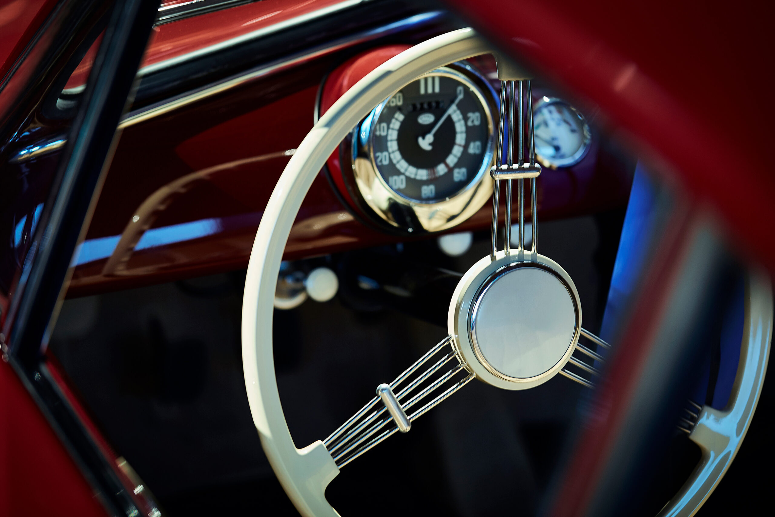 Nahaufnahme des Lenkrads und Armaturenbretts eines Oldtimers mit einem cremefarbenen Lenkrad mit Chrom-Akzenten und einem runden Tachometer mit schwarzen und weißen Markierungen vor einem glänzend roten Innenraum.