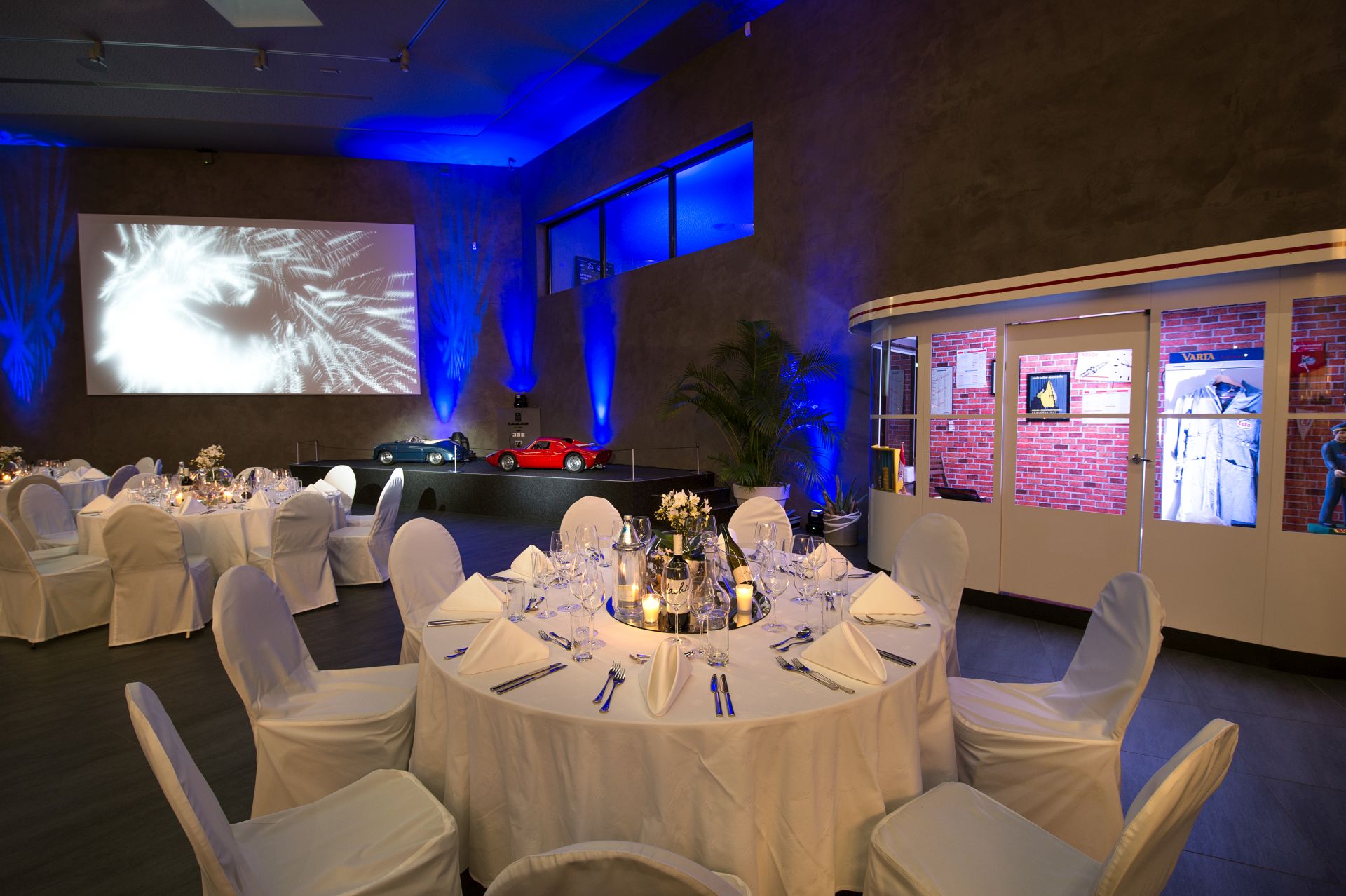 Eleganter Veranstaltungsraum mit runden, weiß gedeckten Tischen für das Abendessen, blauer Stimmungsbeleuchtung, ausgestellten Oldtimern, einem großen Bildschirm mit abstrakten Bildern und einem kleinen Ausstellungsbereich mit Fotos und Erinnerungsstücken.