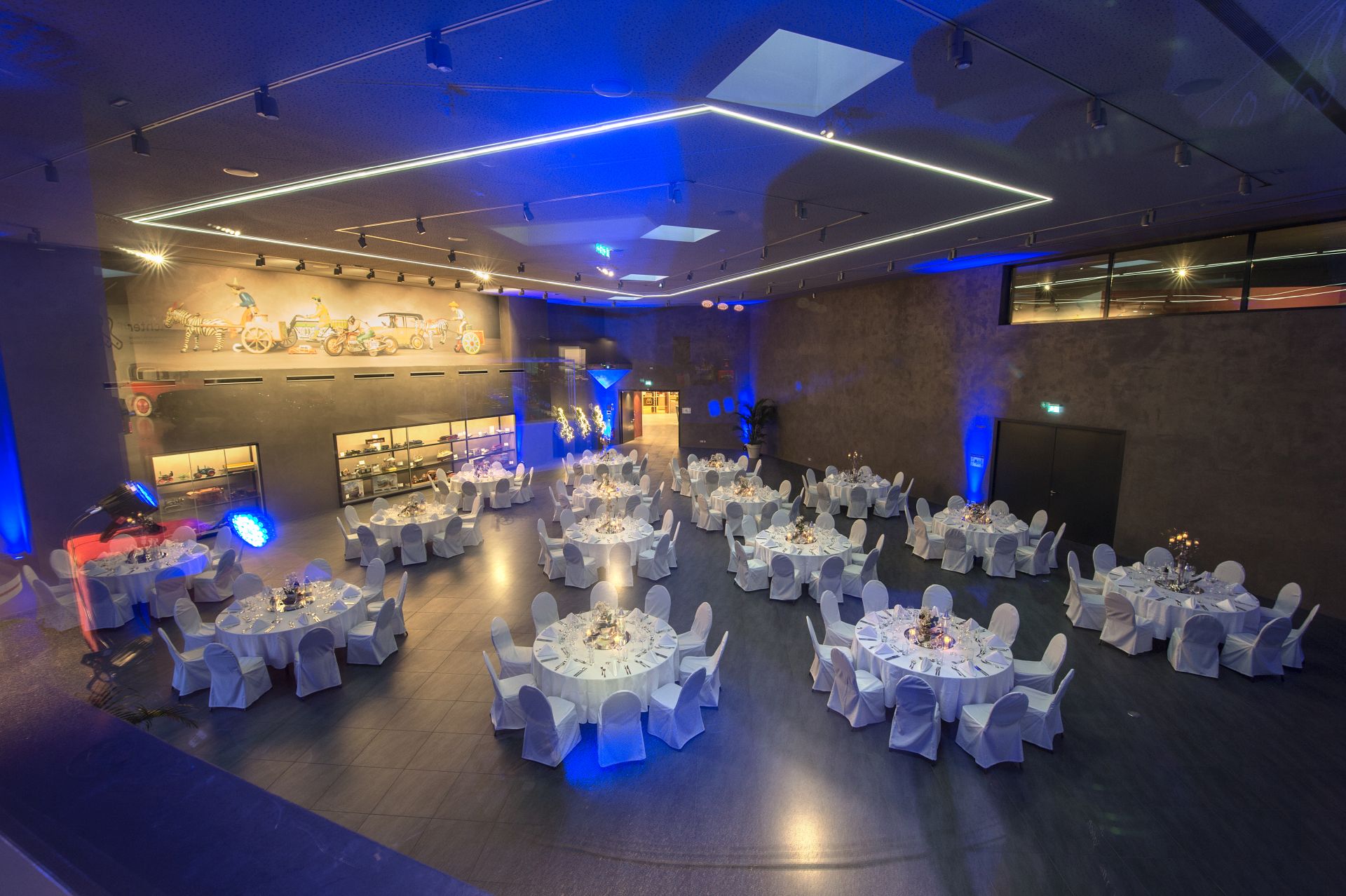 Ein moderner Veranstaltungssaal mit runden, weiß gedeckten Tischen und Stühlen für ein formelles Abendessen, beleuchtet durch blaue Akzente und Wanddisplays, die verschiedene Artikel präsentieren.