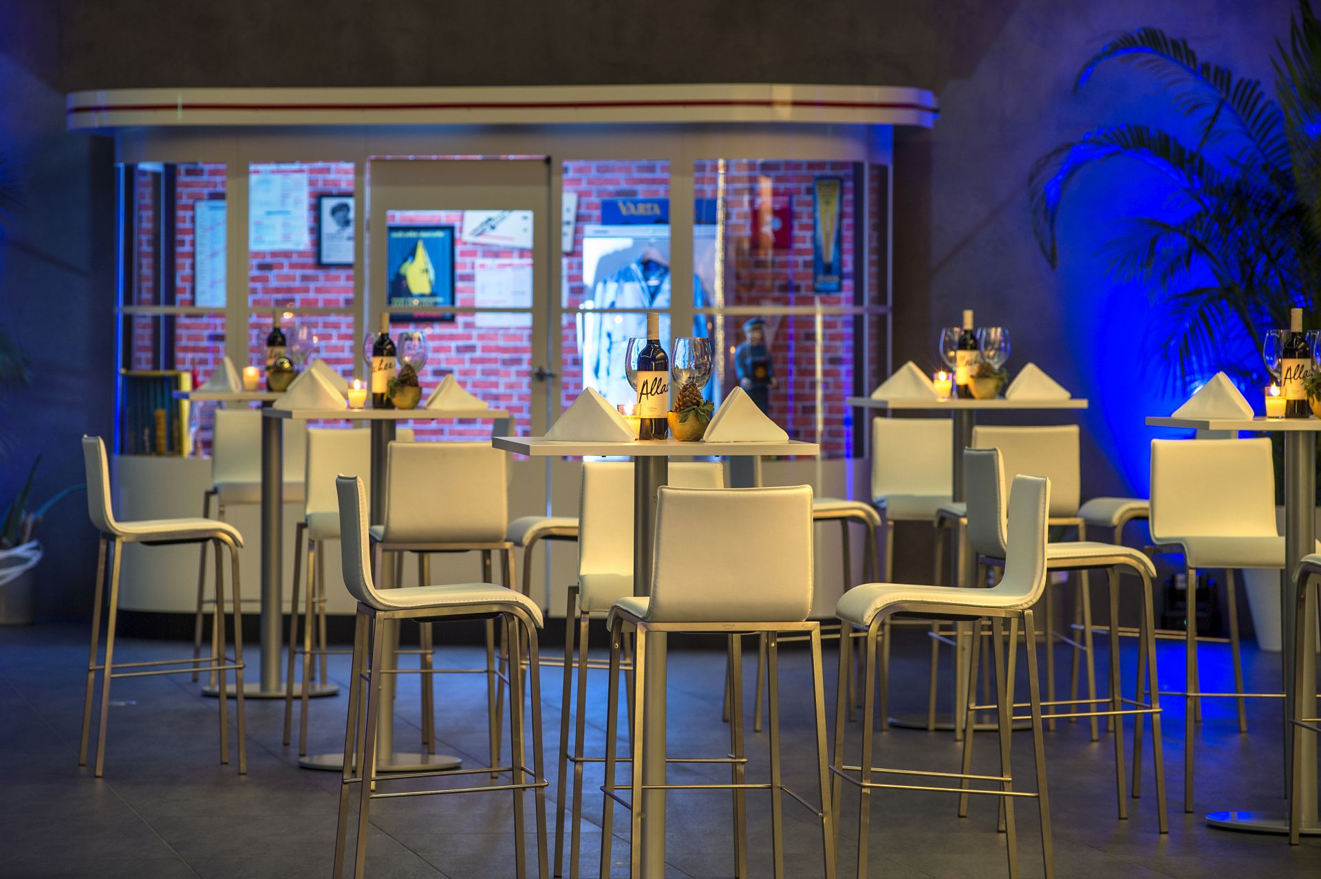 Ein modernes Indoor-Café mit Stehtischen und weißen Barhockern, bestückt mit Gläsern, Servietten und Kerzen. Der Hintergrund besteht aus Plakaten, großen Fenstern, blauer Akzentbeleuchtung und Pflanzen an den Seiten - perfekt als Ausflugsziel in der Nähe von Traumwerk.