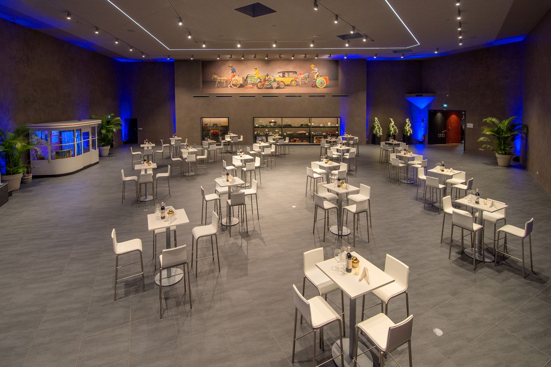 Geräumiger, moderner Speisesaal im Traumwerk mit hoher Decke, grauem Fliesenboden und weit auseinander stehenden weißen Tischen und Stühlen. Wandmalereien, Vitrinen im hinteren Bereich, Topfpflanzen und blaue Lichtakzente schaffen ein perfektes Ausflugsziel.