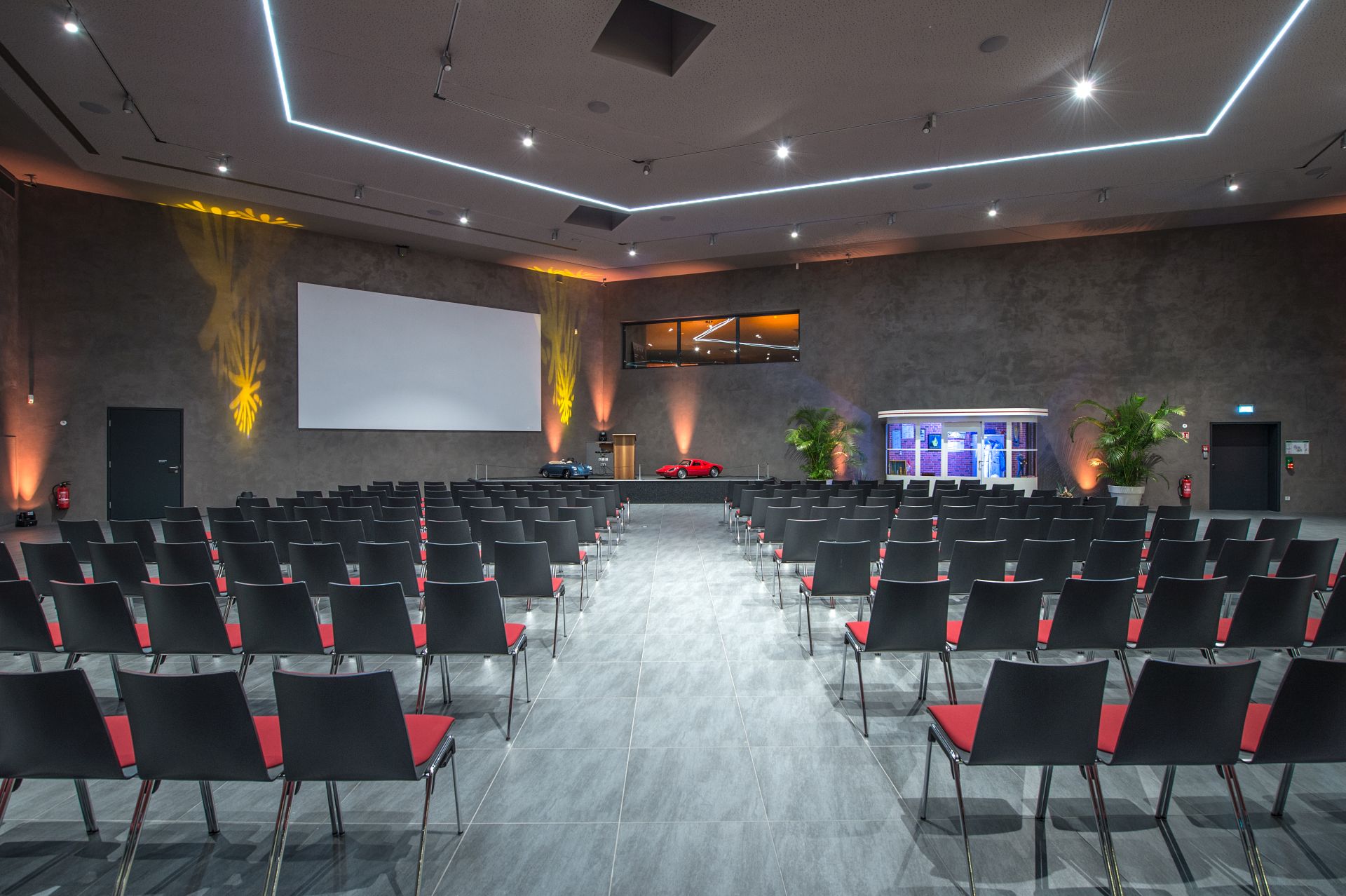Ein moderner Konferenzraum mit schwarzen Stuhlreihen vor einer Bühne, einem großen leeren Bildschirm und zwei Porsche an der Stirnseite. Der Raum ist mit grauen Wänden, Deckenleuchten und Topfpflanzen ausgestattet - perfekt für ein Traumwerk-Ausflugsziel.