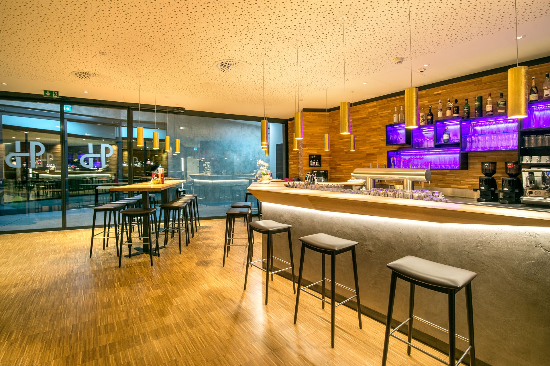 Moderne Bar mit Holzfußboden, hohen Tischen und Hockern, einem gut beleuchteten Tresen, Regalen mit Flaschen und Gläsern und violetter Akzentbeleuchtung. Glastüren führen zu einem weiteren Sitzbereich. Eine warme, einladende Atmosphäre macht es zu einem tollen Ausflugsziel.