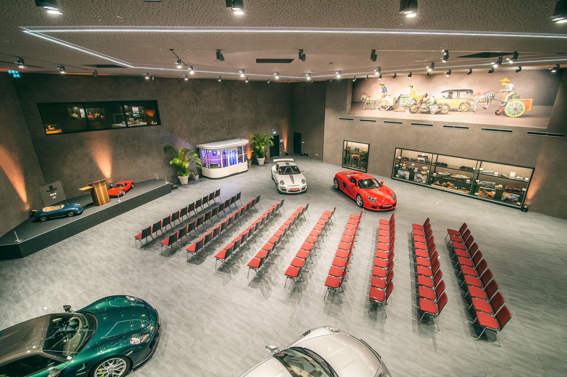 Ein geräumiger Autosalon mit Reihen roter Stühle vor einer kleinen Bühne, mehreren im Raum verteilten Luxusautos und Oldtimer-Kunstwerken an der Wand.
