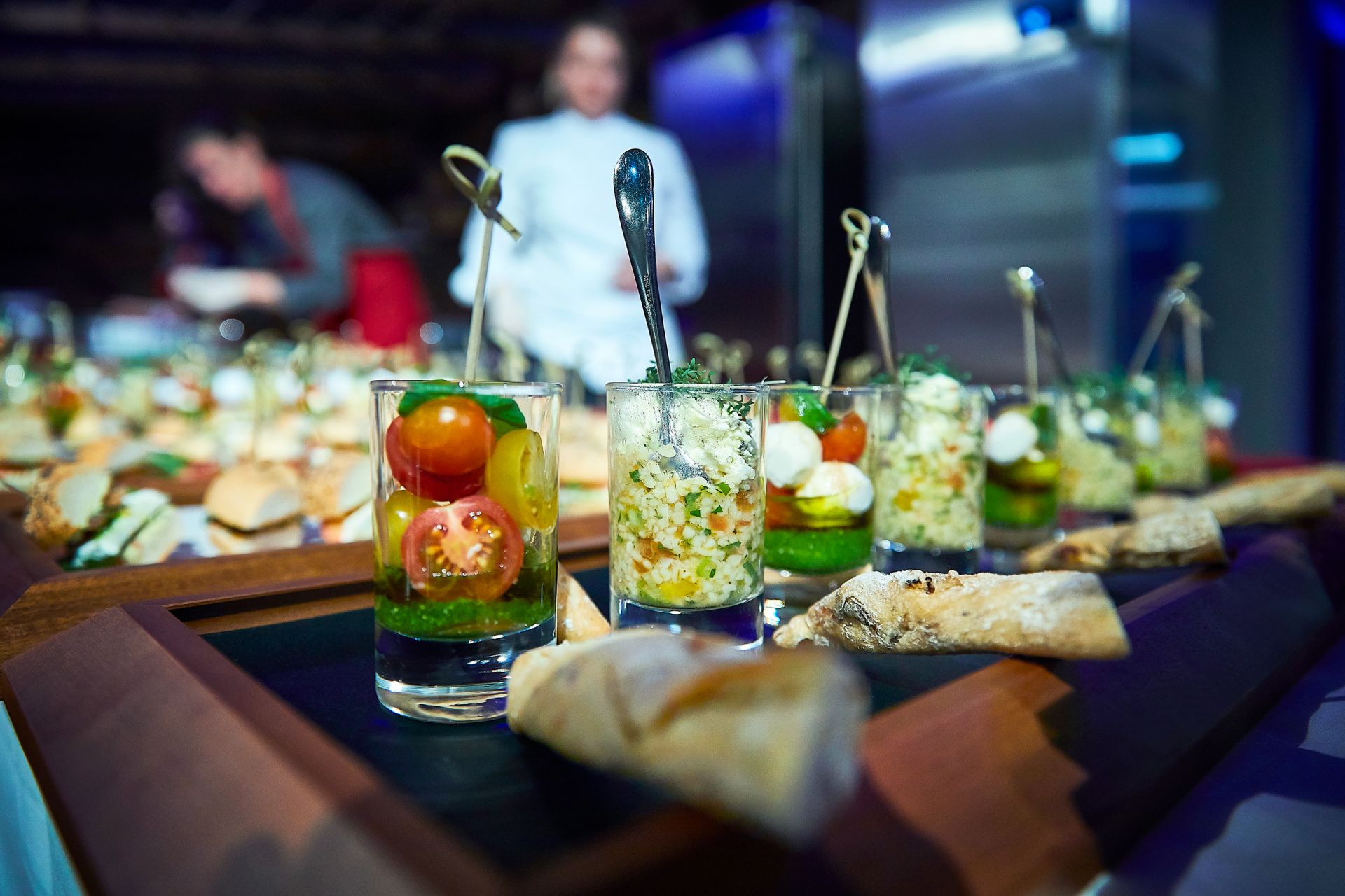 Nahaufnahme von Gourmet-Häppchen in Glasbechern mit geschichtetem Gemüse, garniert mit Kräutern und Spießen, arrangiert auf einem Tablett mit Brotstücken - perfekt für Besucher des Traumwerks, einem beliebten Ausflugsziel. Die Köche sind im Hintergrund verschwommen.