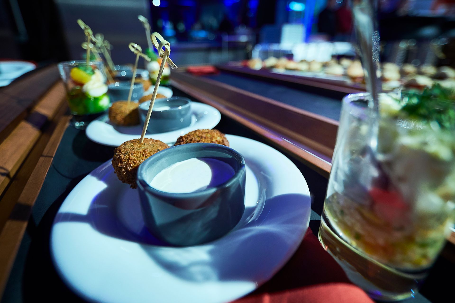 Eine Nahaufnahme einer Reihe weißer Teller mit Vorspeisen, darunter runde, gebratene Häppchen am Spieß und Dip-Sauce, auf einem Buffet im Traumwerk, mit anderen kleinen Gerichten im Hintergrund.