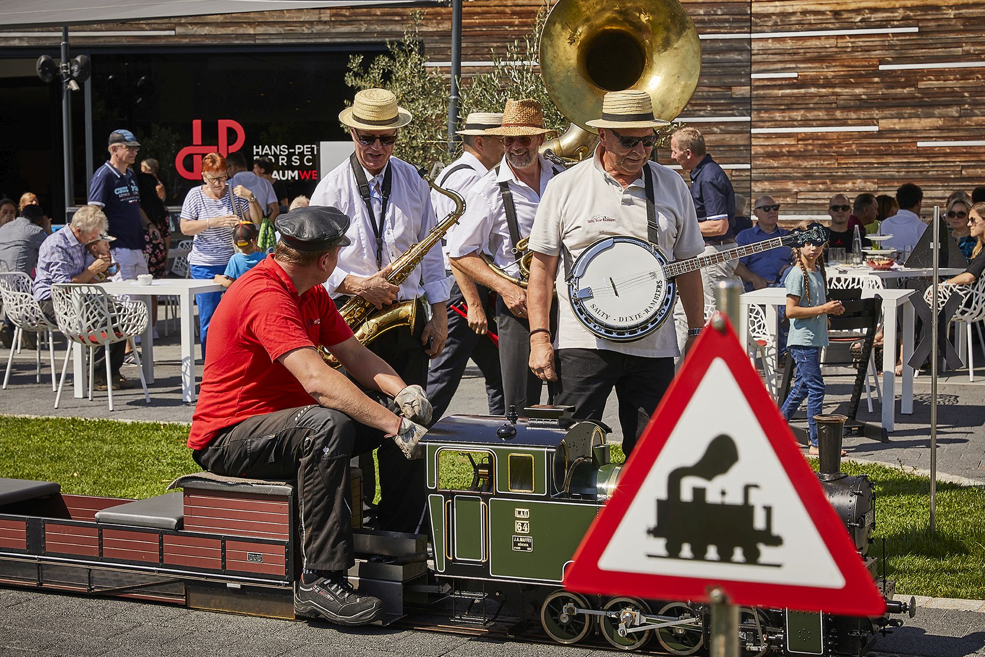 Eine kleine Band mit Saxophon, Banjo und Sousaphon spielt im Freien in der Nähe eines Cafés am beliebten Ausflugsziel Traumwerk, während ein Mann in einem roten Hemd eine Modelleisenbahn an einem Bahnübergangsschild vorbeifährt. Im Hintergrund sitzen Menschen an Tischen.